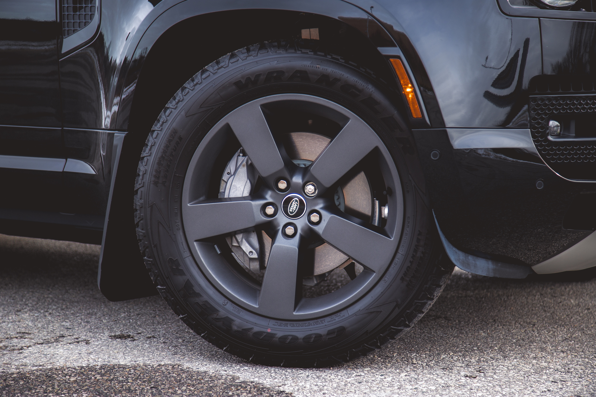 Close-up image of the wheel and tire of a Land Rover Defender, featuring a matte black alloy rim and Goodyear Wrangler tire. The reflective surface of the vehicle's paint is visible in the background.