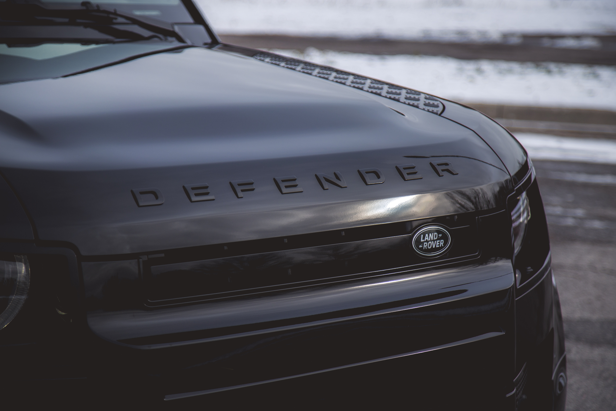 Close-up view of the front grille and hood of a black 2026 Land Rover Defender, featuring the embossed 'DEFENDER' logo and the Land Rover emblem.