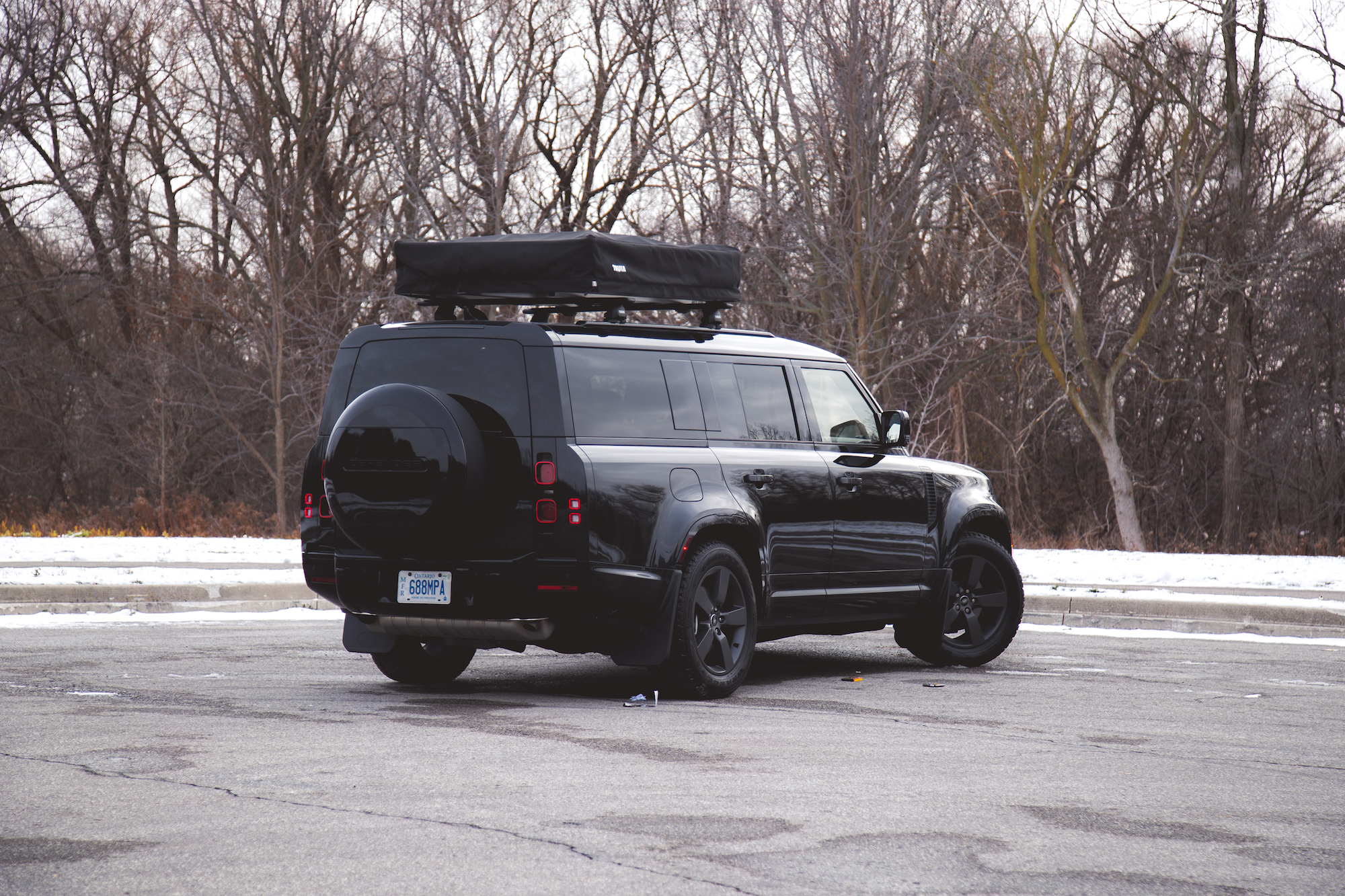 A black Land Rover Defender parked with a rooftop tent attached, surrounded by bare trees in a winter landscape.
