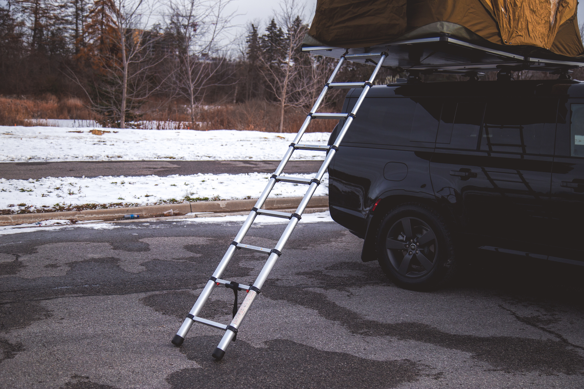 An aluminum ladder leaning against a black 2026 Land Rover Defender 130 with a tent mounted on the roof, set against a background of snow and bare trees.