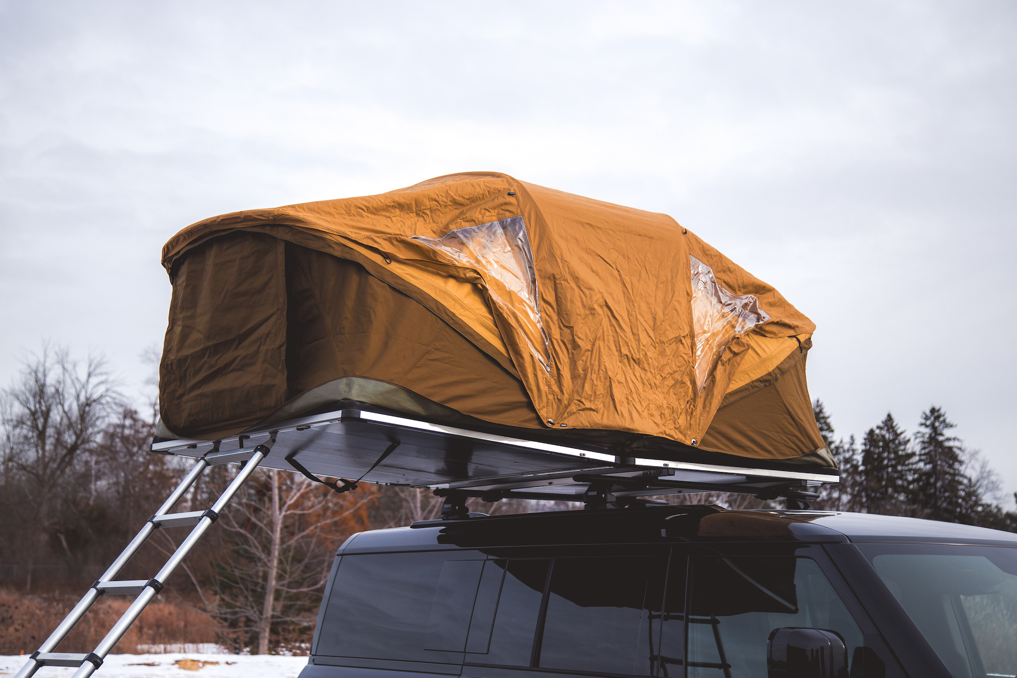 A rooftop tent in mustard yellow is mounted on the roof of a black SUV, with a ladder extending down to the ground. The tent has visible windows allowing light inside, set against a cloudy sky.