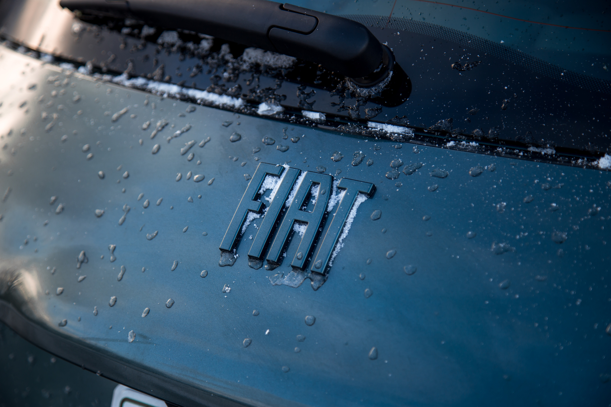 Close-up of a wet Fiat logo on a car's rear hatch, with droplets of water and snow visible.