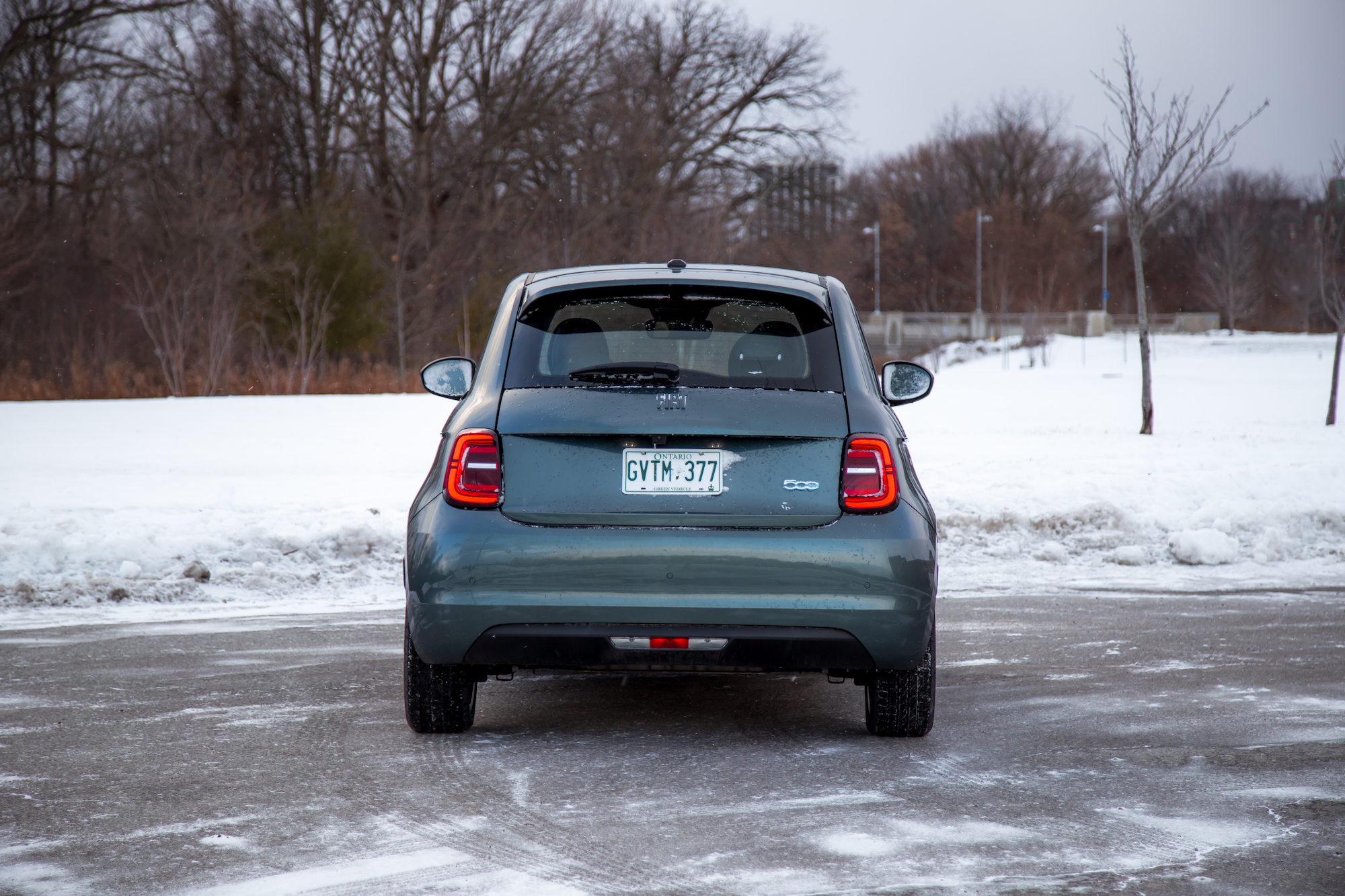 A rear view of a grey compact car parked on a snowy surface, with trees in the background and a cloudy sky.