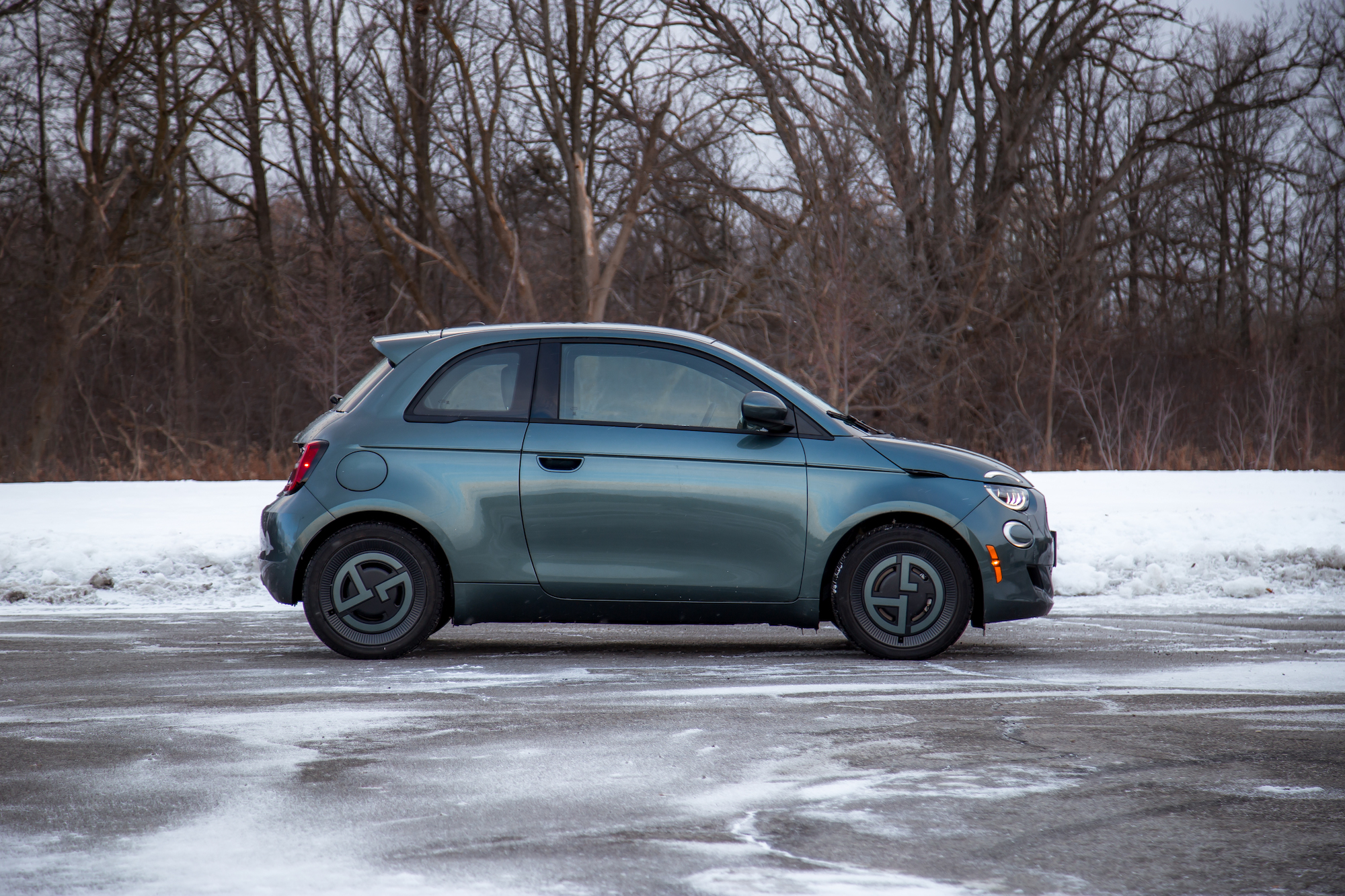 A compact electric car parked on a snow-covered surface, showcasing its side profile against a background of bare trees.