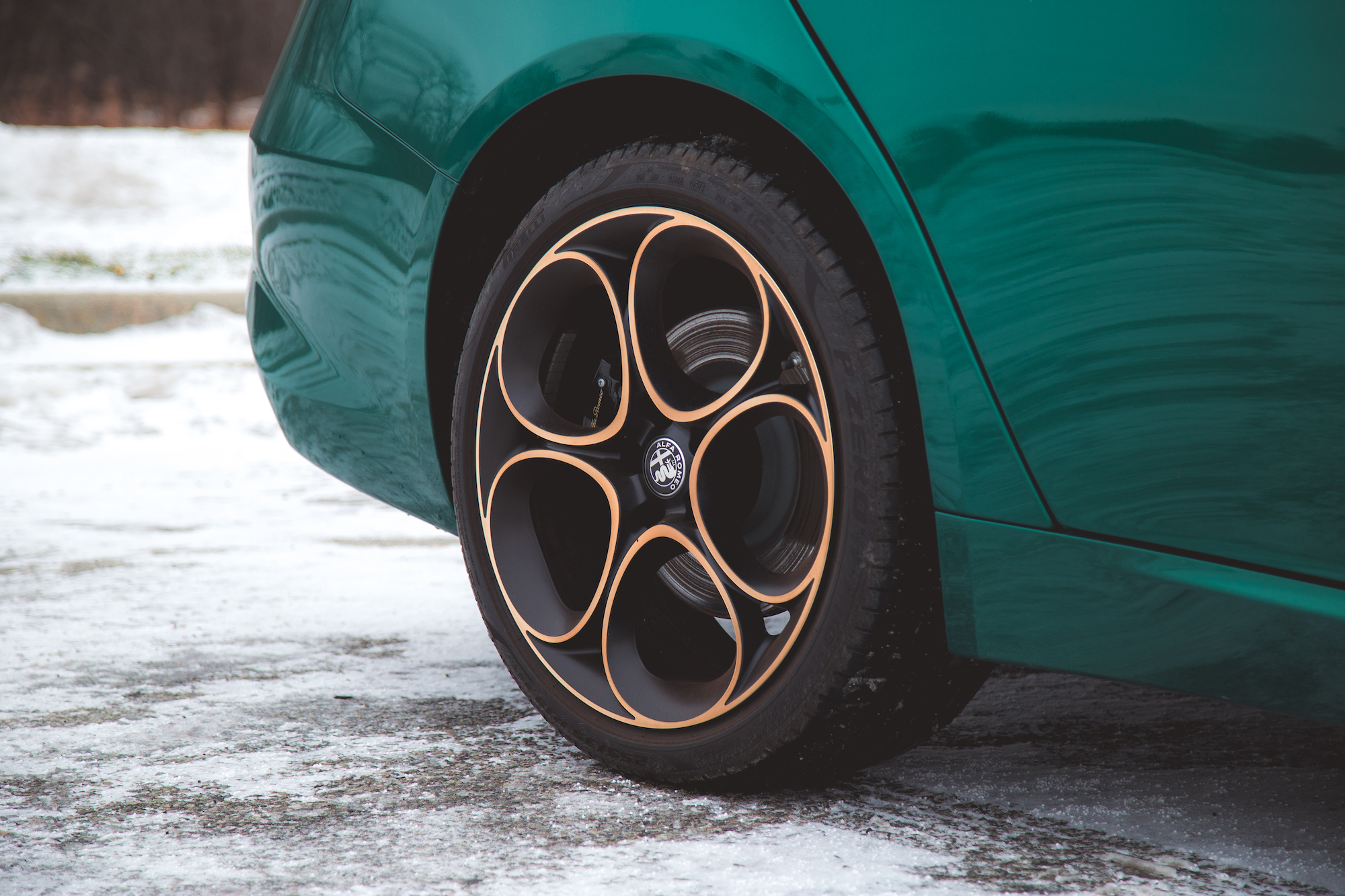 Primer plano de una rueda de automóvil única con un borde negro y dorado, sobre un fondo nevado con una carrocería verde parcialmente visible.