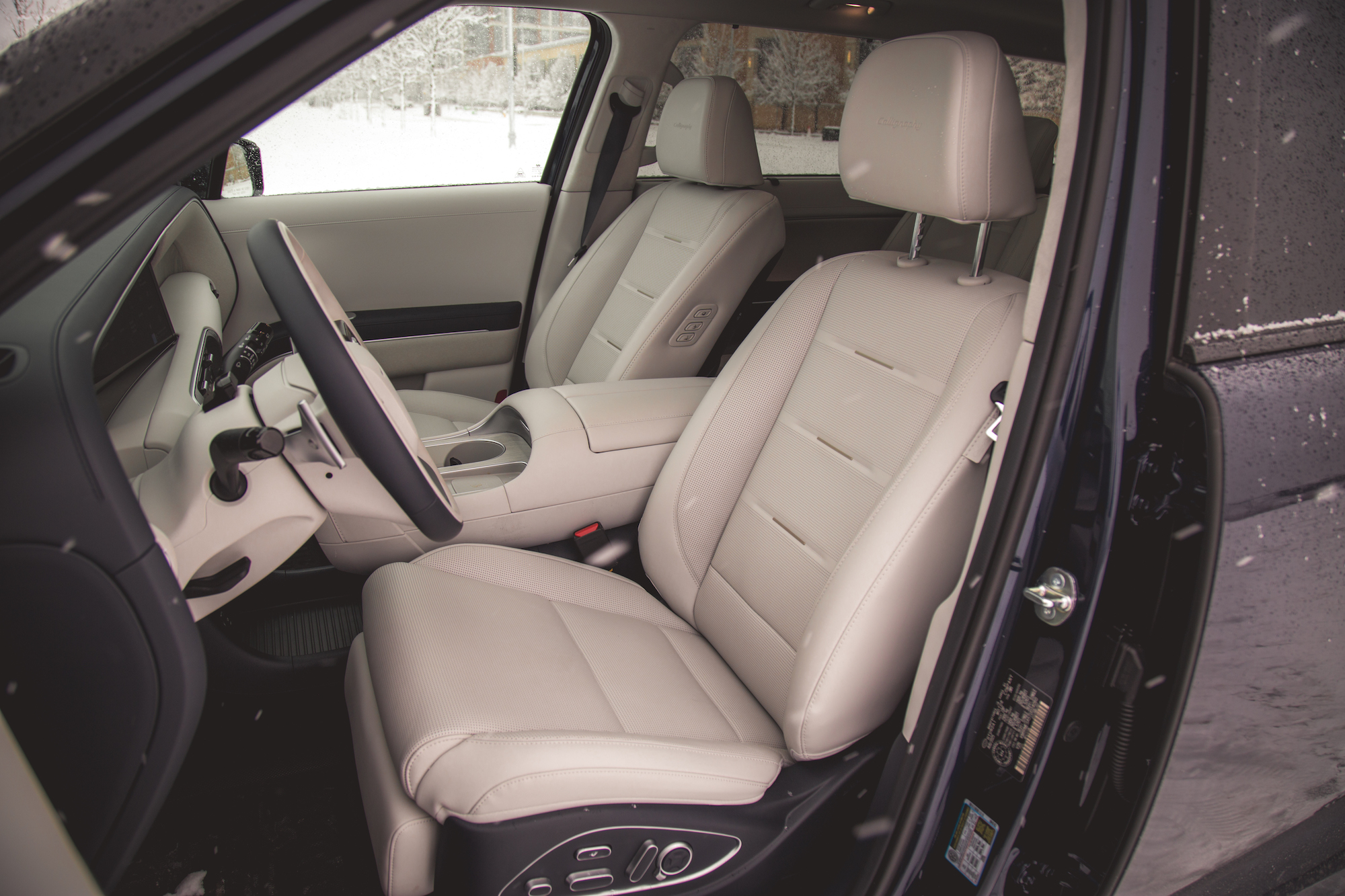 Vista interior de un coche que muestra dos asientos de cuero beige y un volante, con un paisaje nevado visible a través de la ventanilla.