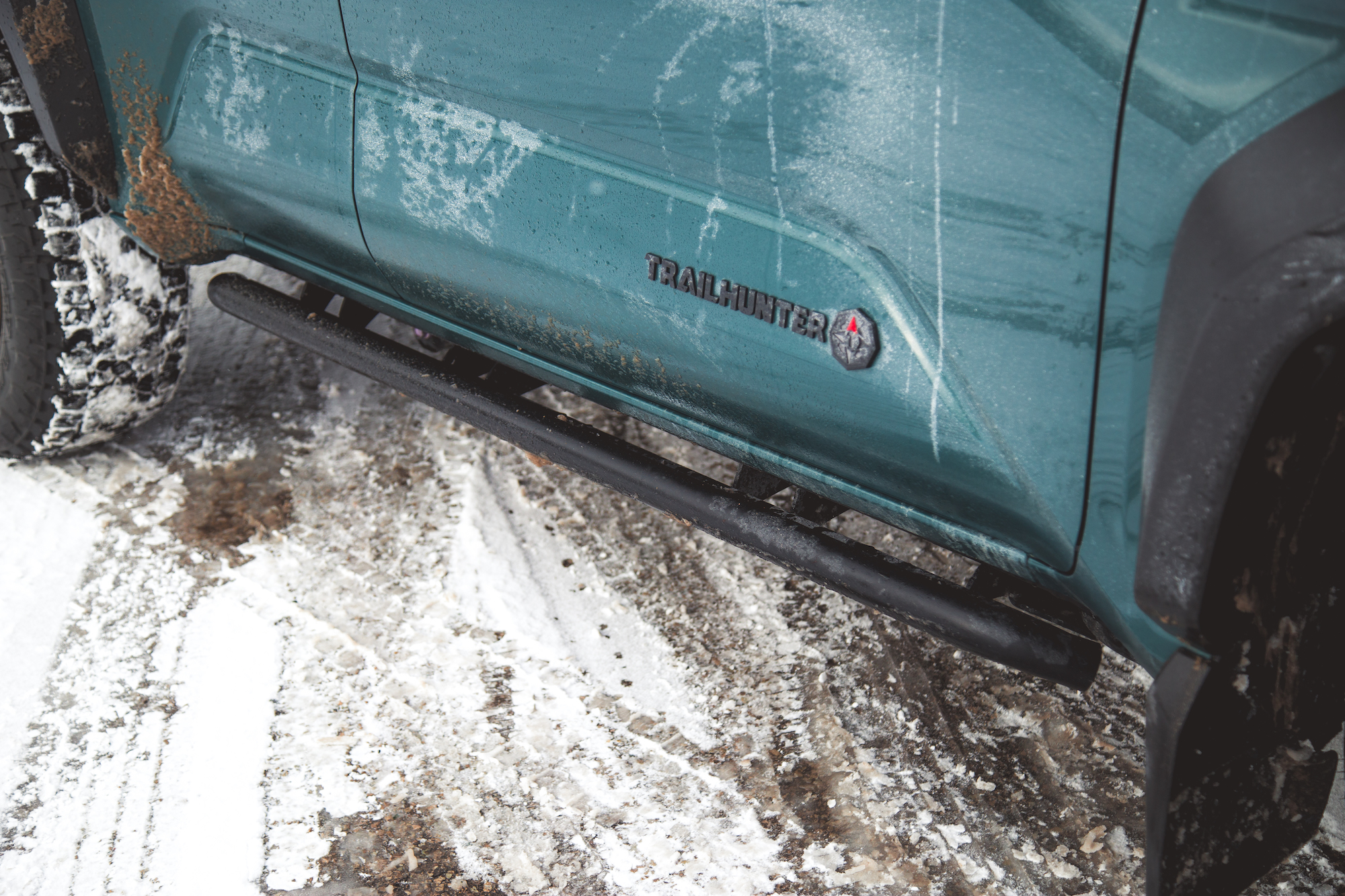 Primer plano del costado de un vehículo verde azulado que muestra la insignia Trailhunter y un escalón lateral negro, con terreno nevado al fondo.