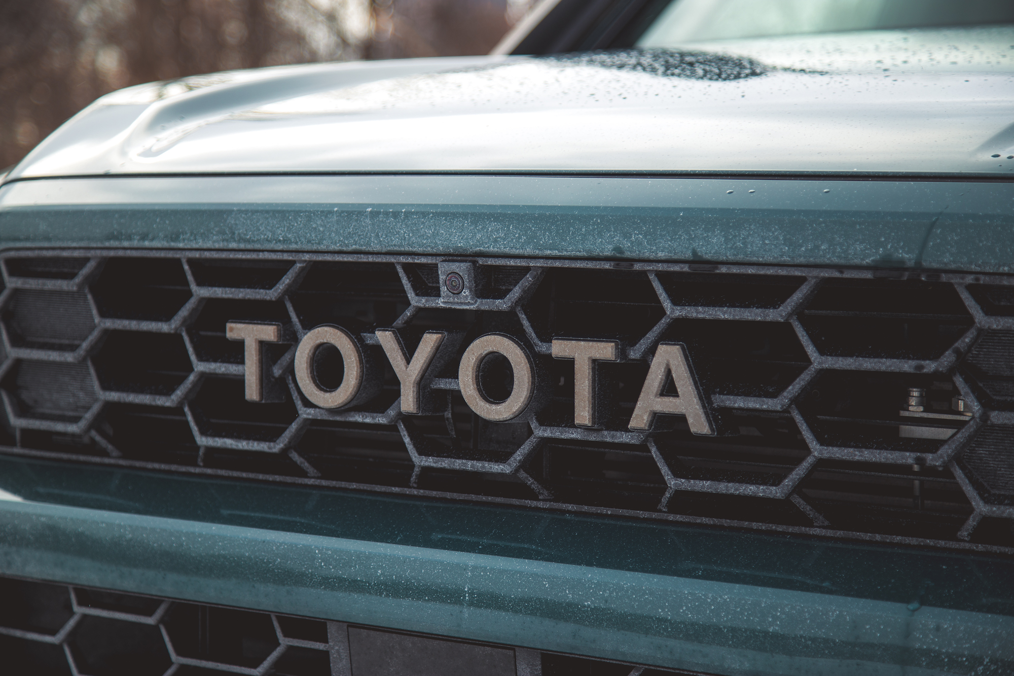 Primer plano de la parrilla delantera de un vehículo Toyota que muestra el logotipo y una cámara, con un acabado verde y una superficie ligeramente polvorienta.