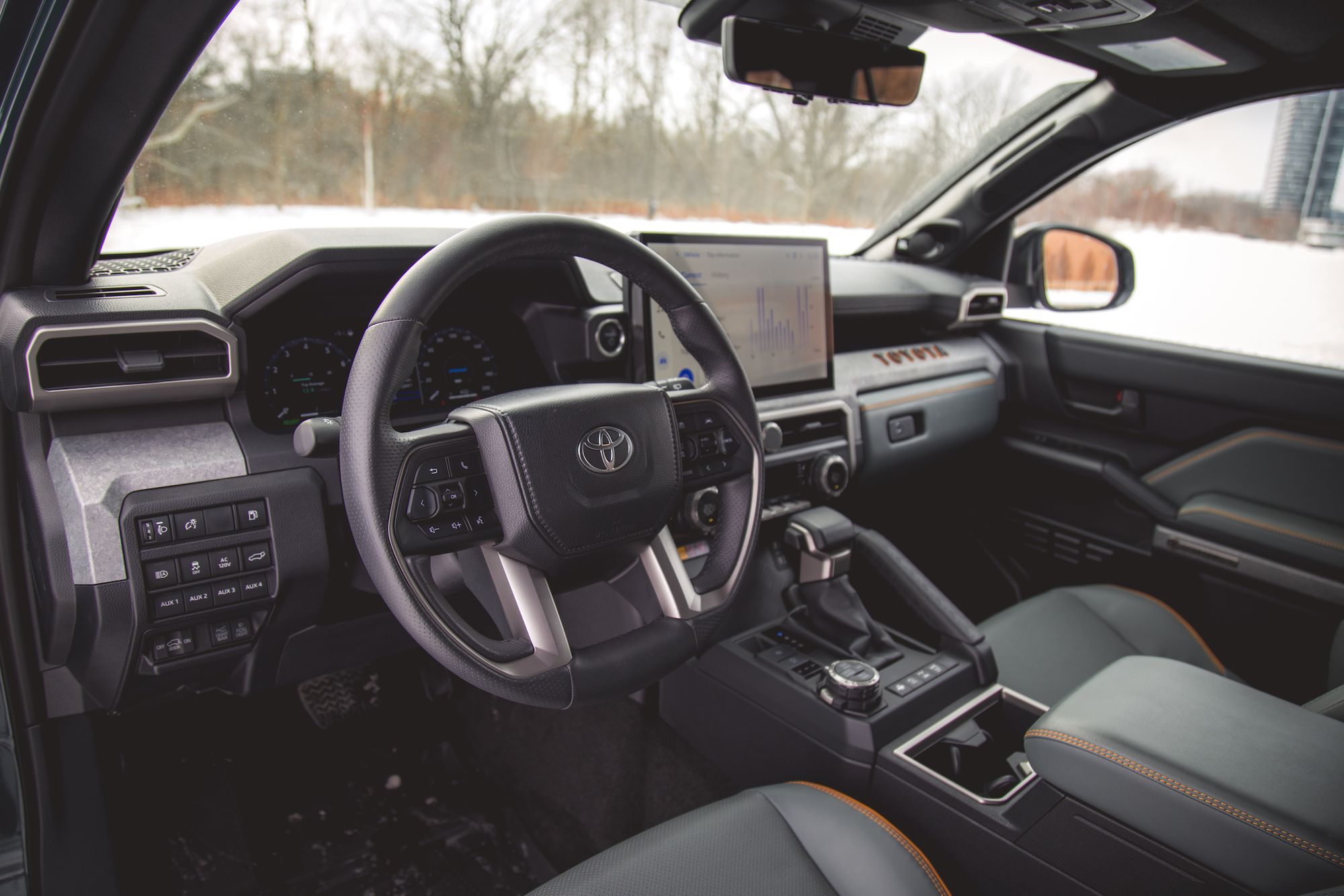 Vista interior de un vehículo Toyota moderno con volante de cuero, tablero digital y una gran pantalla de información y entretenimiento. Los asientos son grises con costuras naranjas y la cabina está diseñada para una estética contemporánea.