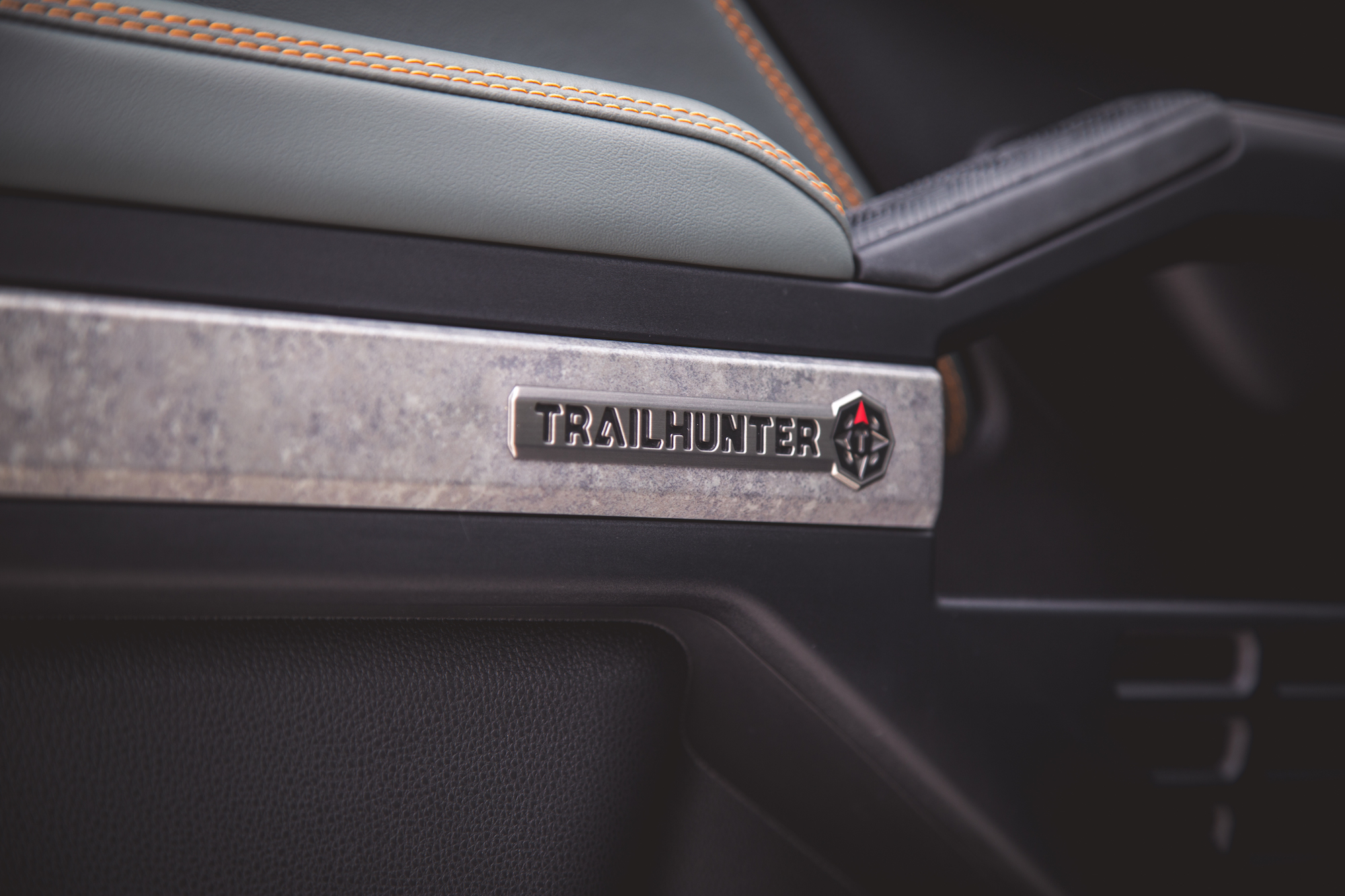 Primer plano del interior de un automóvil con una insignia 'TRAILHUNTER' en un panel elegante con detalles texturizados y costuras naranjas.