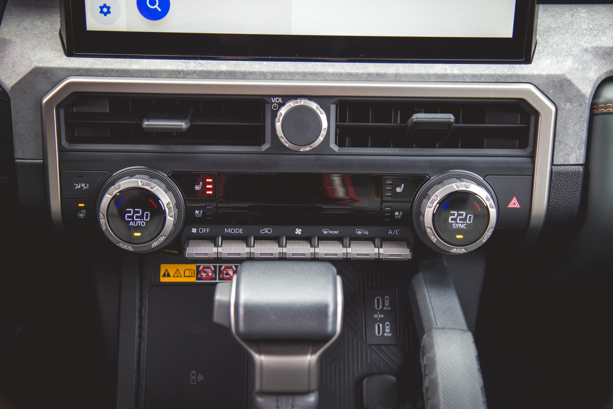 Primer plano del tablero de un automóvil moderno con perillas de control de clima, una pantalla táctil y varios controles que incluyen configuraciones de volumen y aire acondicionado.