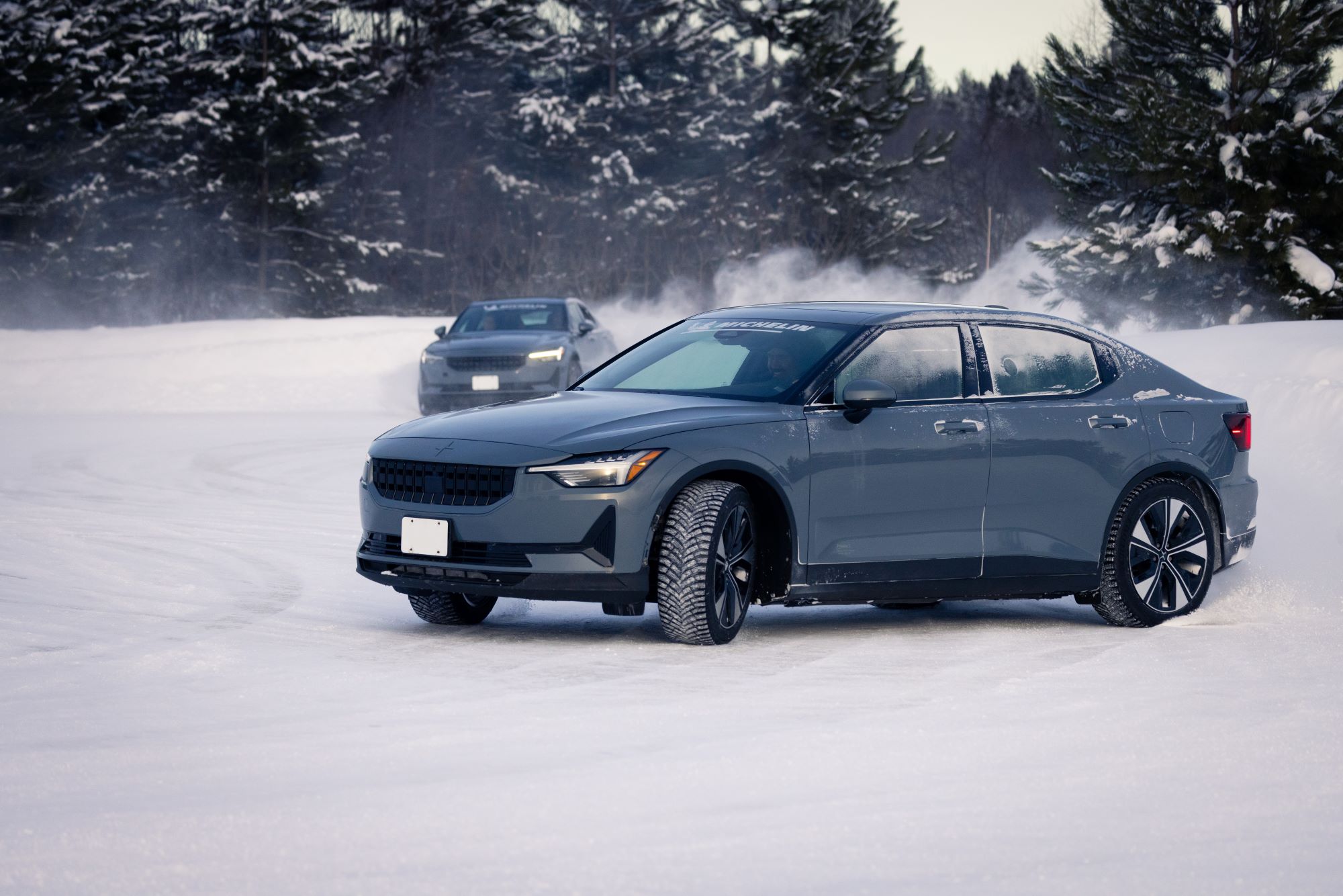 A gray sedan driving in a snowy landscape, showcasing winter traction, with another vehicle visible in the background.