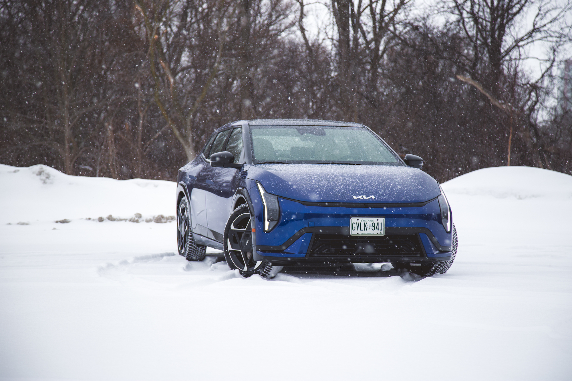 Un vehículo Kia azul estacionado en un paisaje nevado, con copos de nieve cayendo a su alrededor.