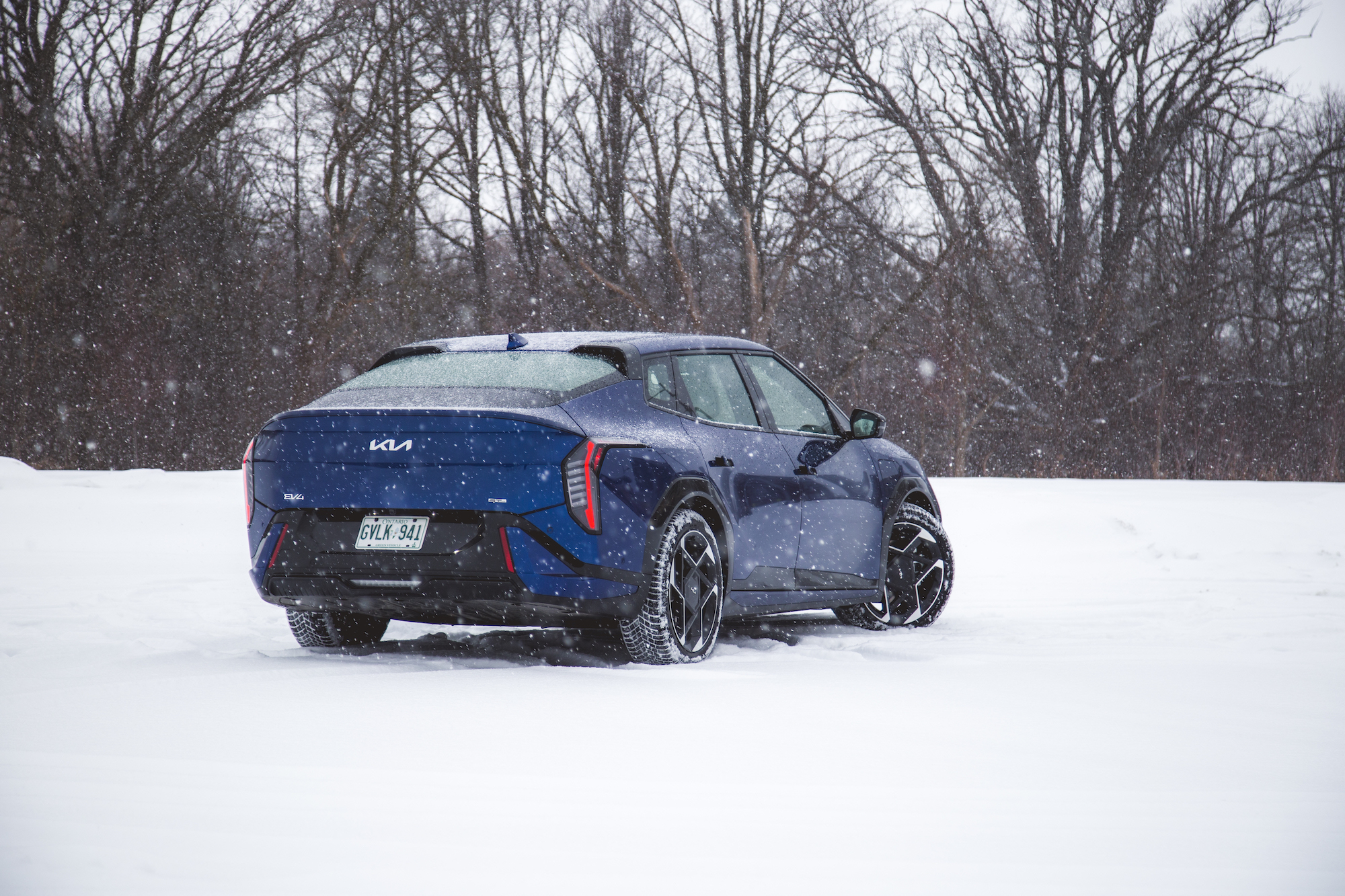 Un coche eléctrico azul estacionado en un paisaje nevado con copos de nieve cayendo y árboles desnudos al fondo.