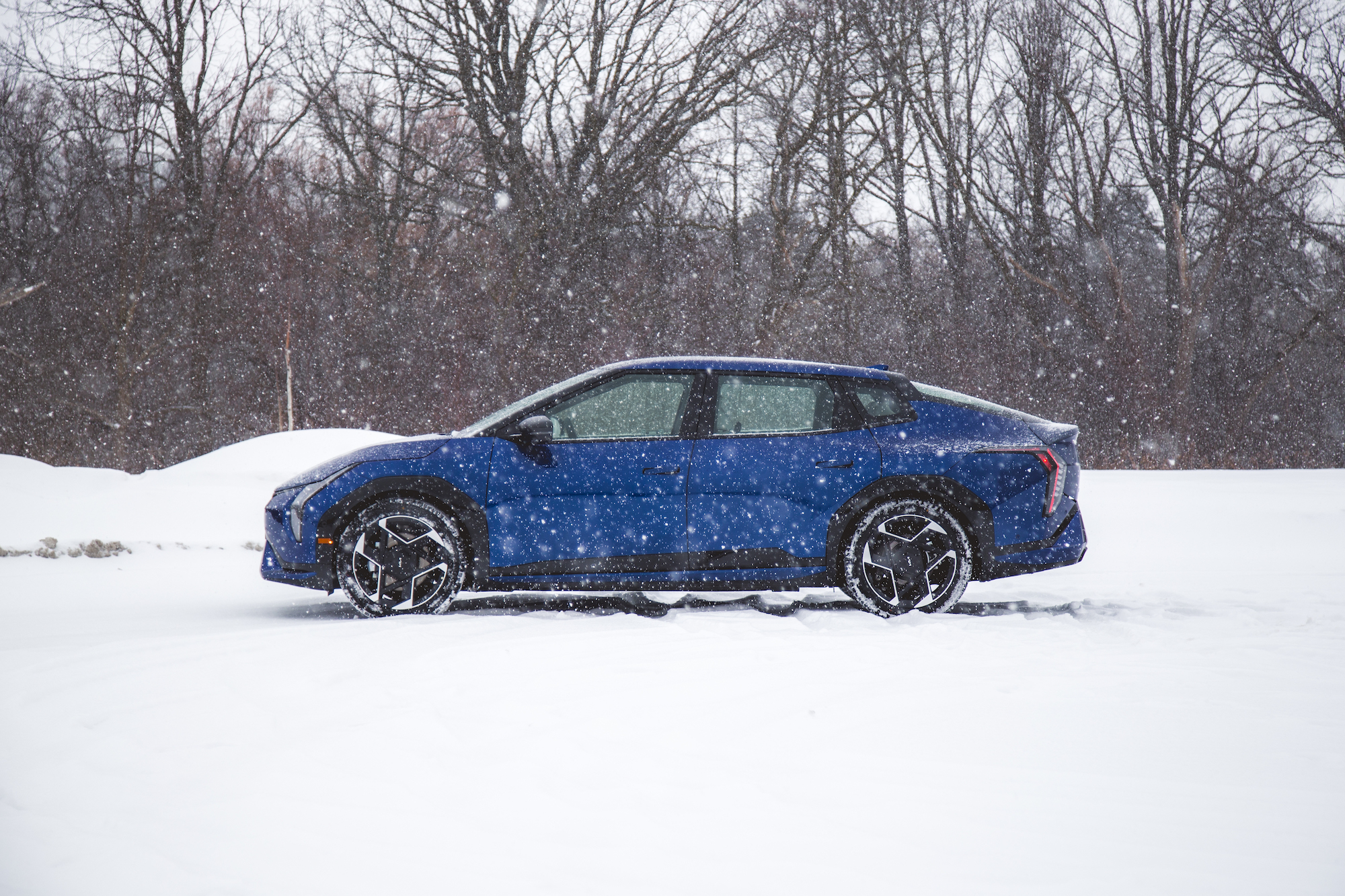 Un coche azul estacionado en un paisaje nevado con copos de nieve cayendo y árboles al fondo.
