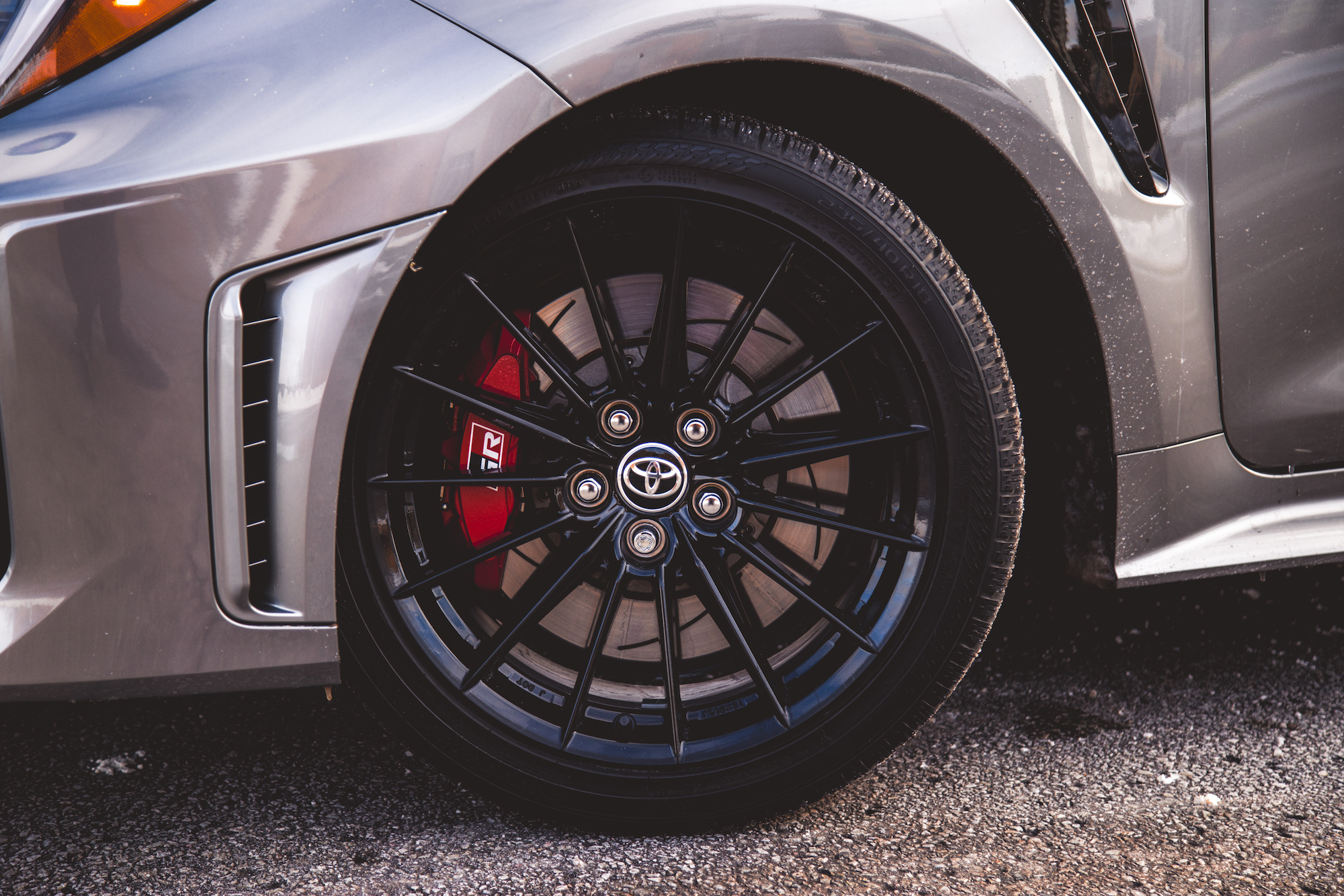 Primer plano de una llanta de aleación negra con una pinza de freno roja con el logotipo de Toyota, montada en un automóvil plateado.