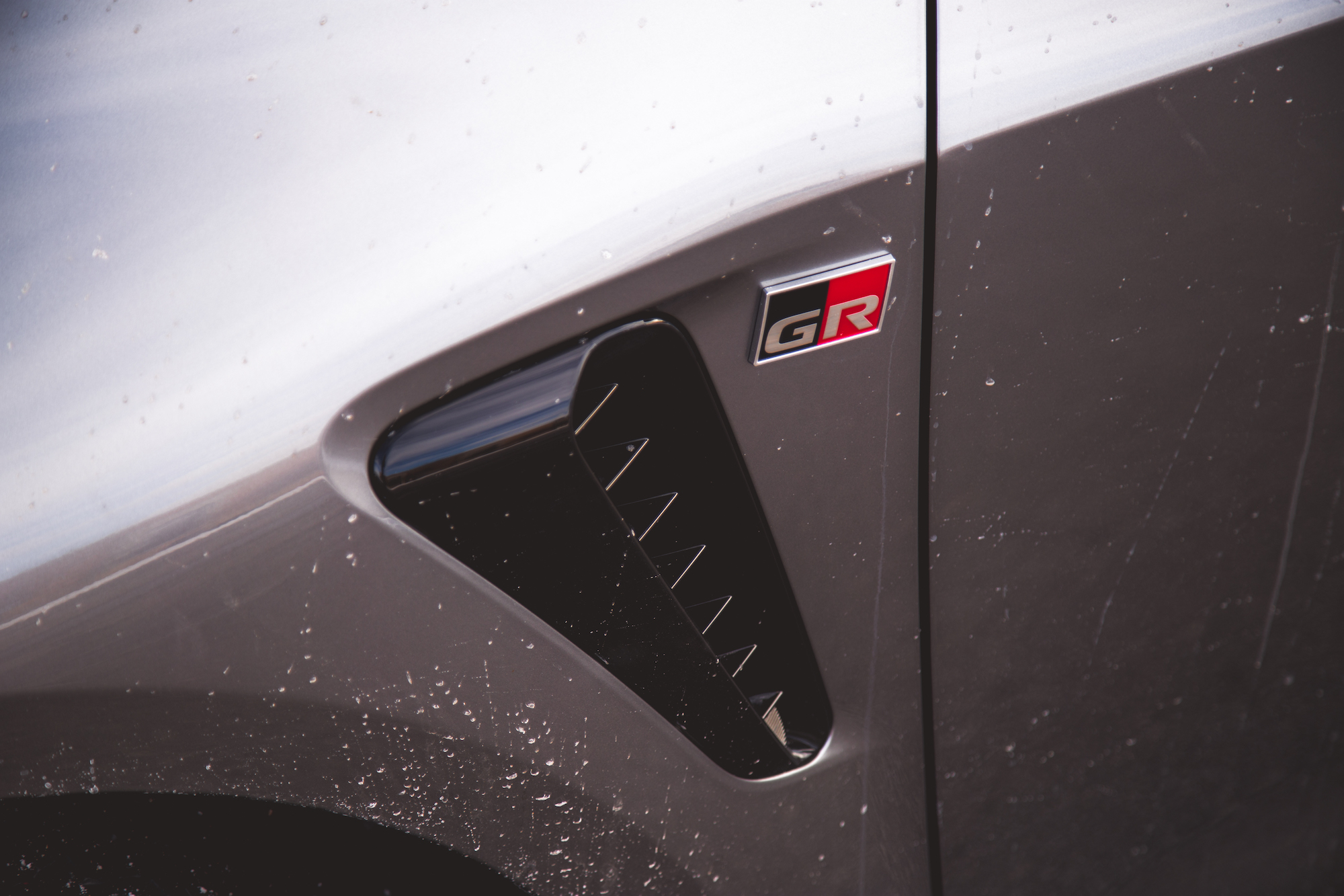 Primer plano del lateral de un automóvil plateado con un emblema GR y un detalle de ventilación negro.