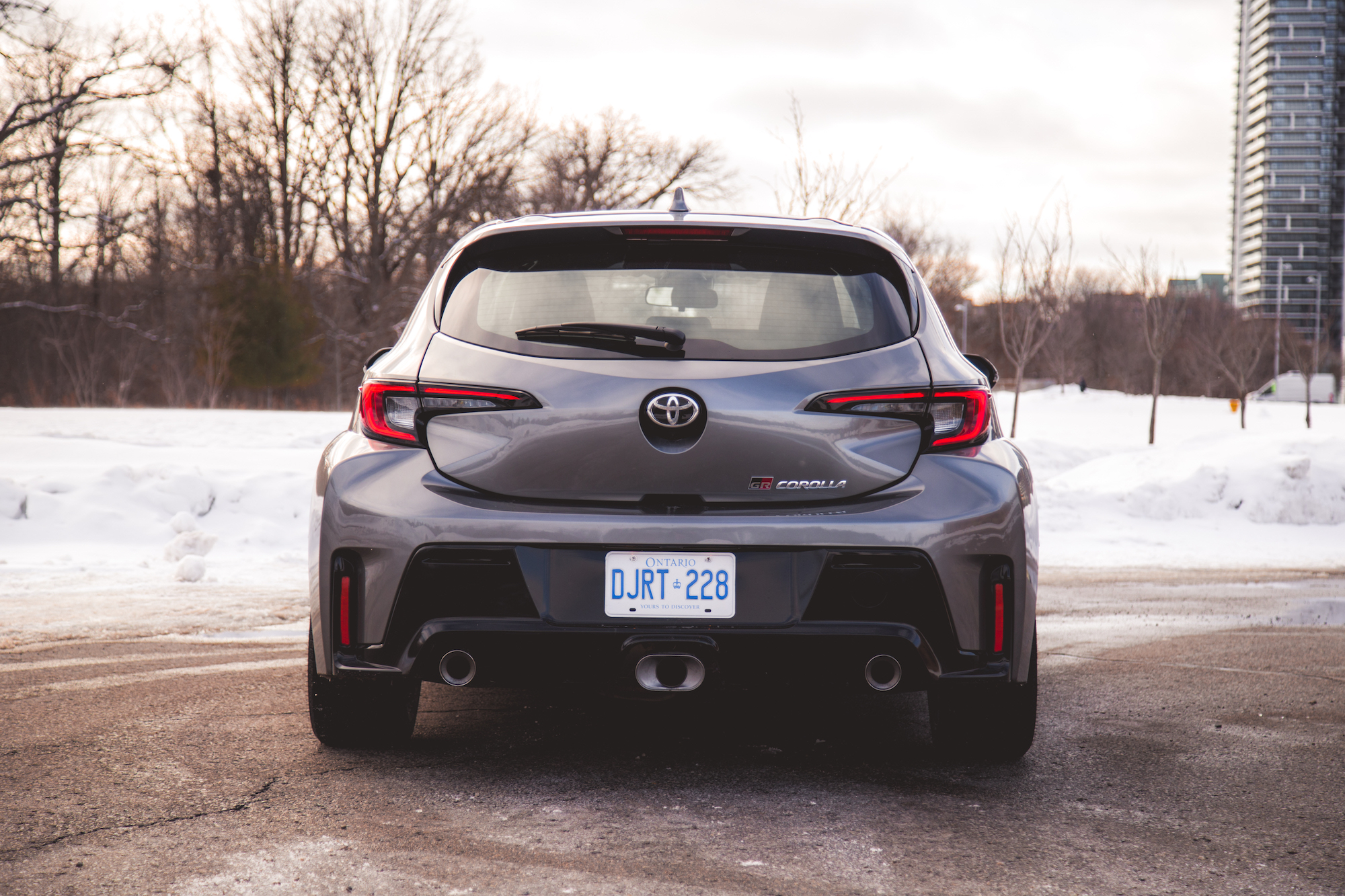 Vista trasera de un Toyota GR Corolla gris estacionado sobre una superficie nevada, con árboles y un edificio al fondo.