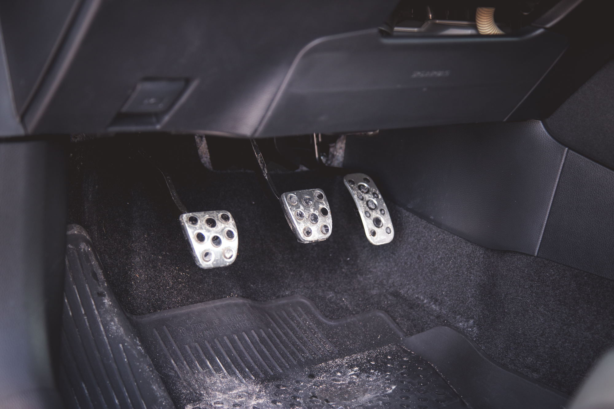 Vista cercana del área de los pedales de un automóvil que muestra tres pedales metálicos sobre una superficie alfombrada negra.