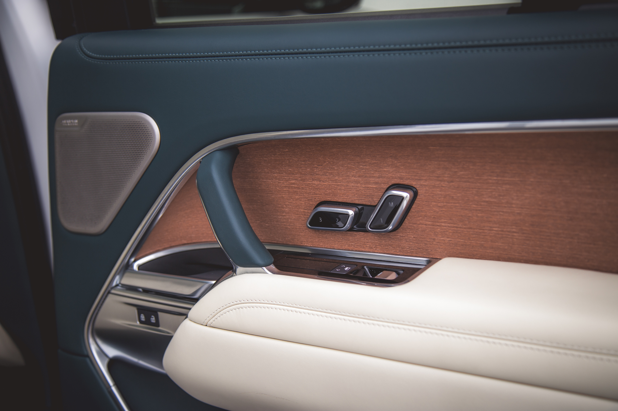 Vista cercana del panel de la puerta de un automóvil de lujo con materiales de cuero y madera, botones de control para ventanas y ajustes de asientos, y una rejilla de altavoz.