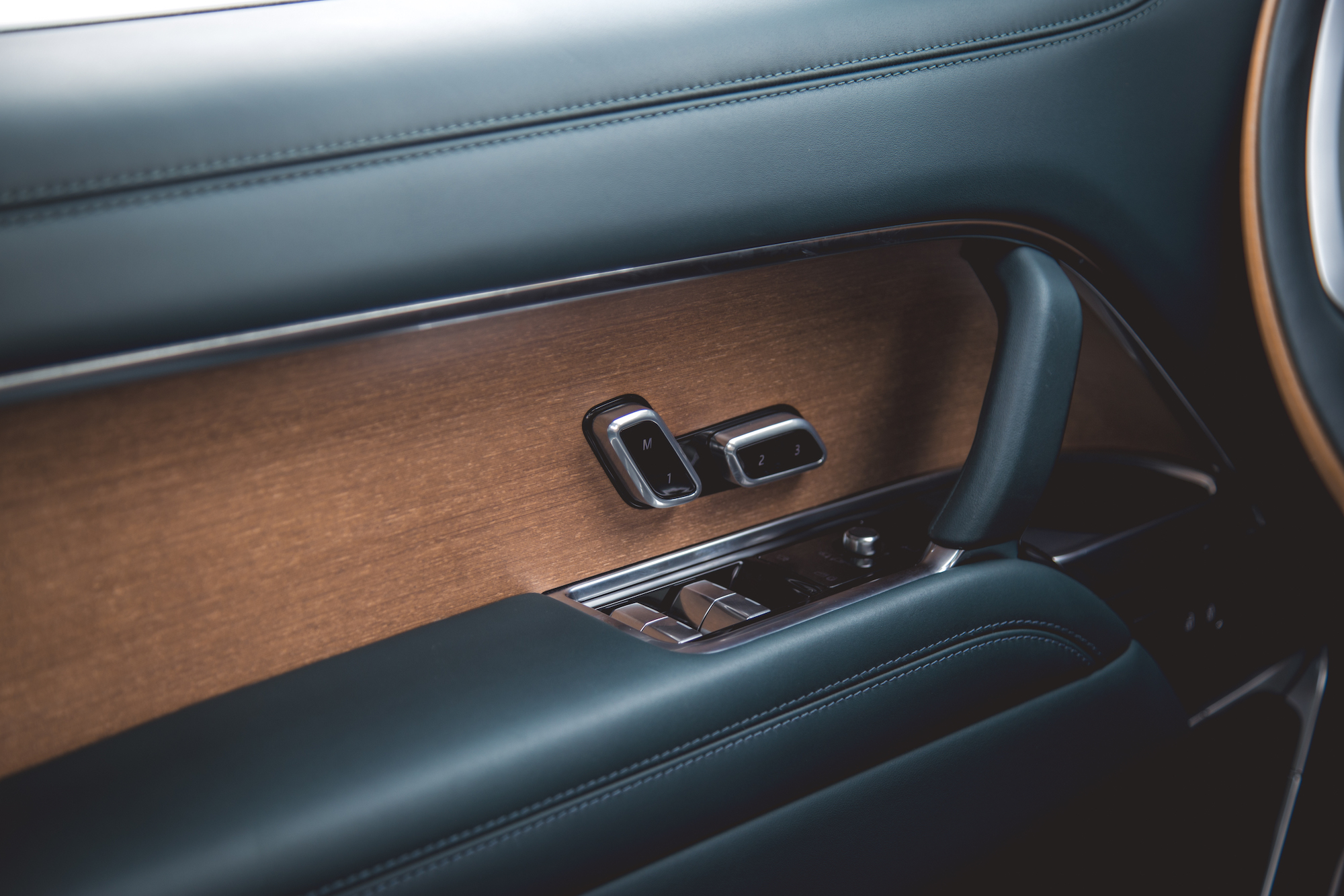 Primer plano del interior de la puerta de un automóvil con molduras de madera, reposabrazos de cuero y botones de control de ventanas.