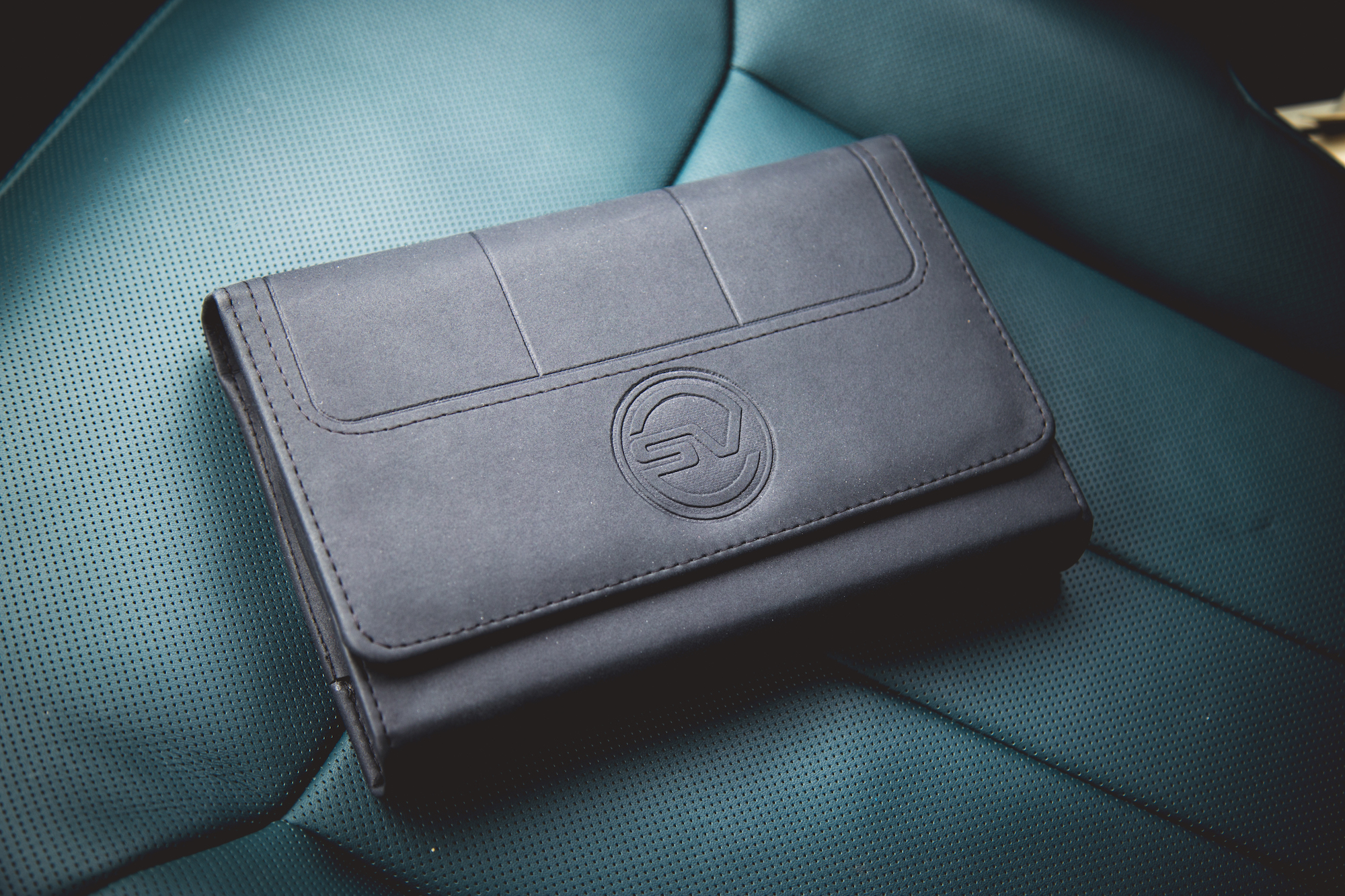Una billetera de cuero negro con un logo, colocada sobre una superficie texturizada en verde azulado.