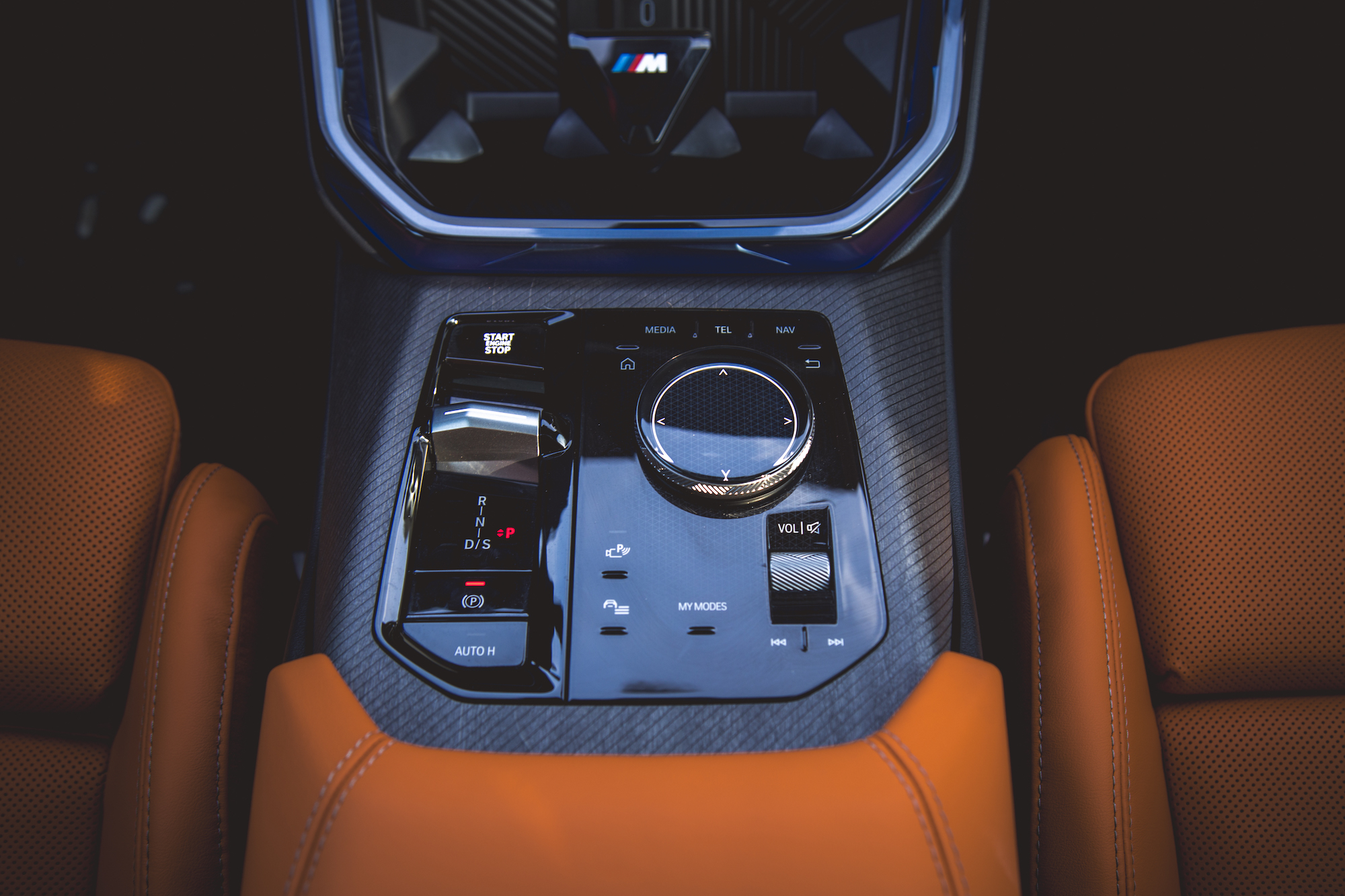 Close-up view of a car's center console featuring a gear selector, start/stop button, and multimedia controls, with orange perforated leather seats visible.