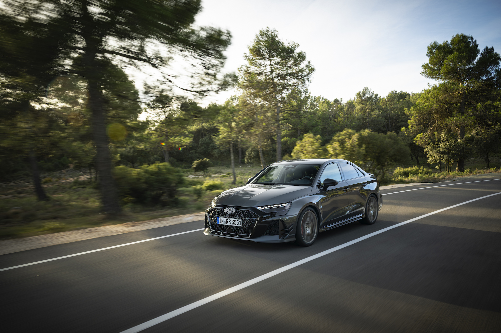 A sleek black car driving on a road surrounded by trees and greenery, showcasing motion with blurred background.