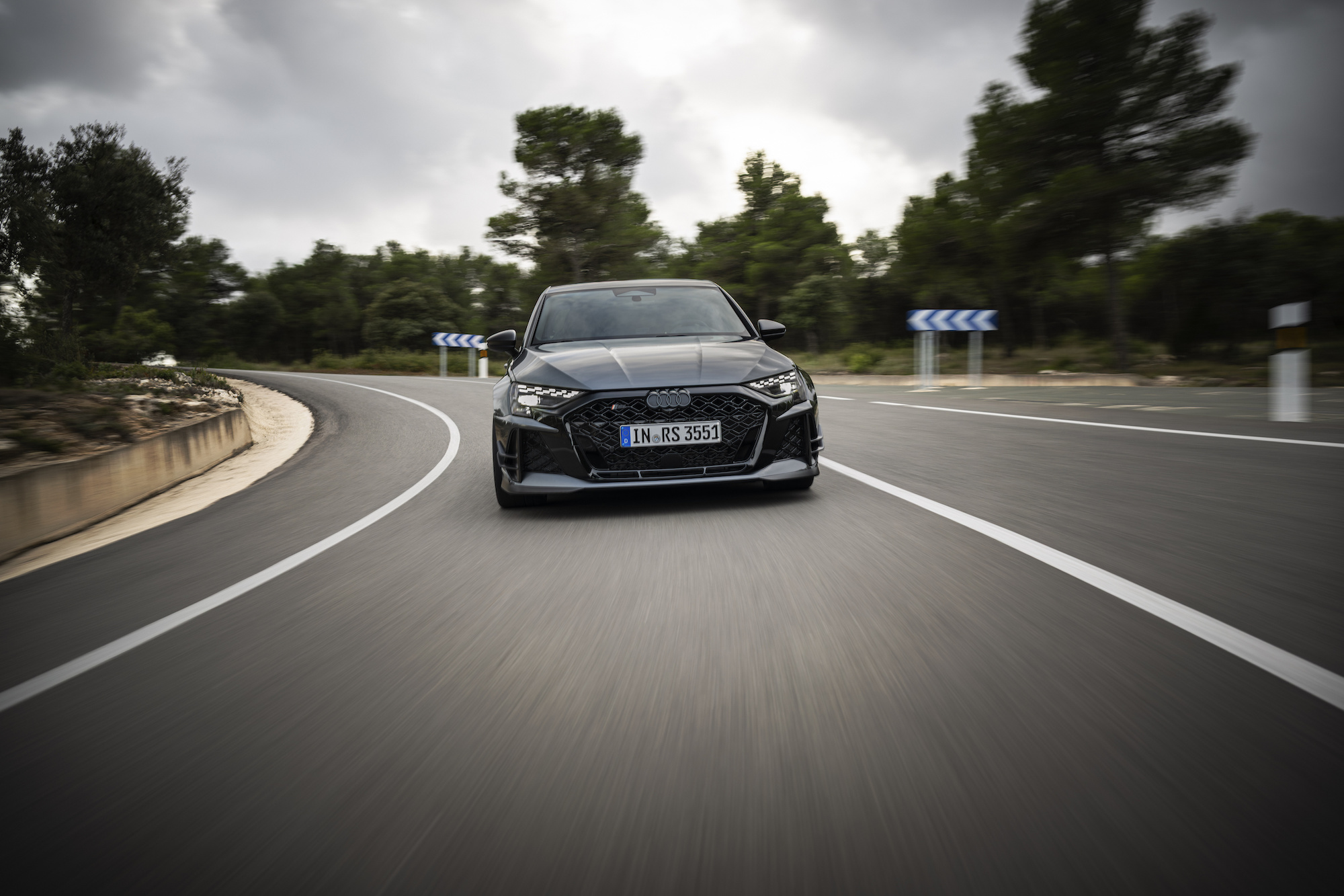A black Audi RS7 sports car speeding around a curve on a rural road, with a backdrop of green trees and overcast skies.