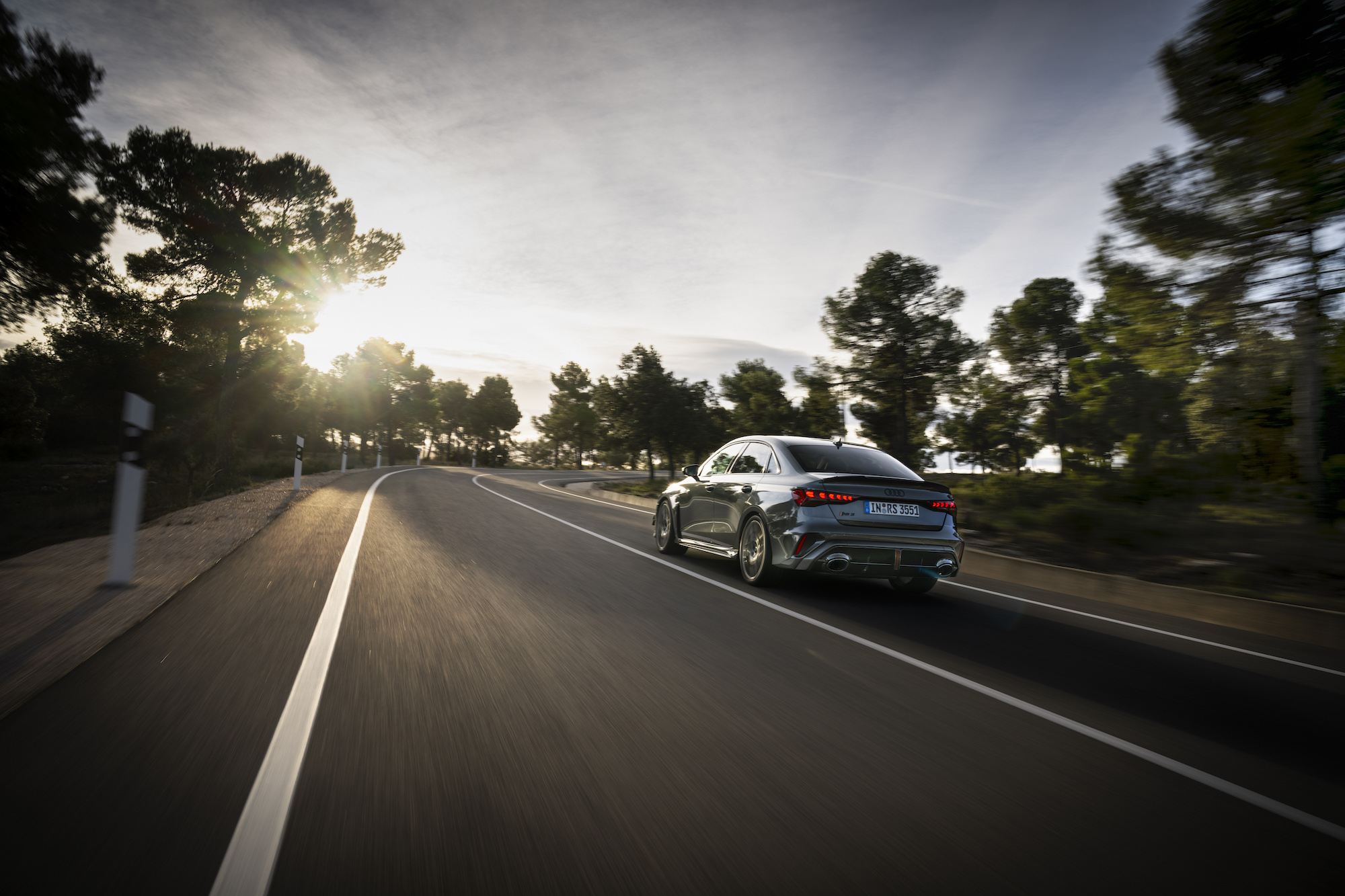A sleek silver car driving on a winding road surrounded by trees, with the sun shining in the background.