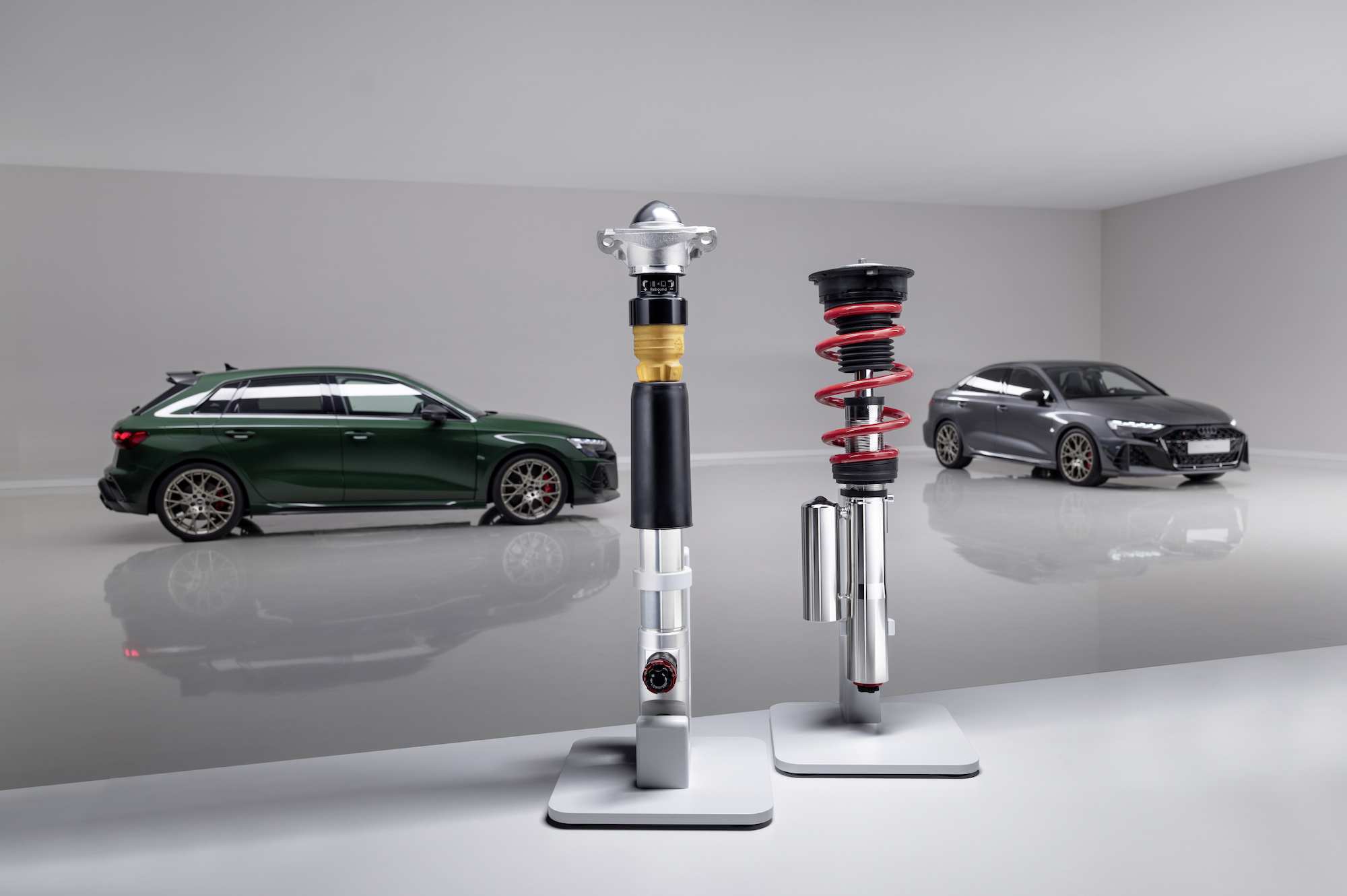 Two high-performance automotive suspension systems in foreground with a dark green sporty car in the background and a grey sporty car on the right.