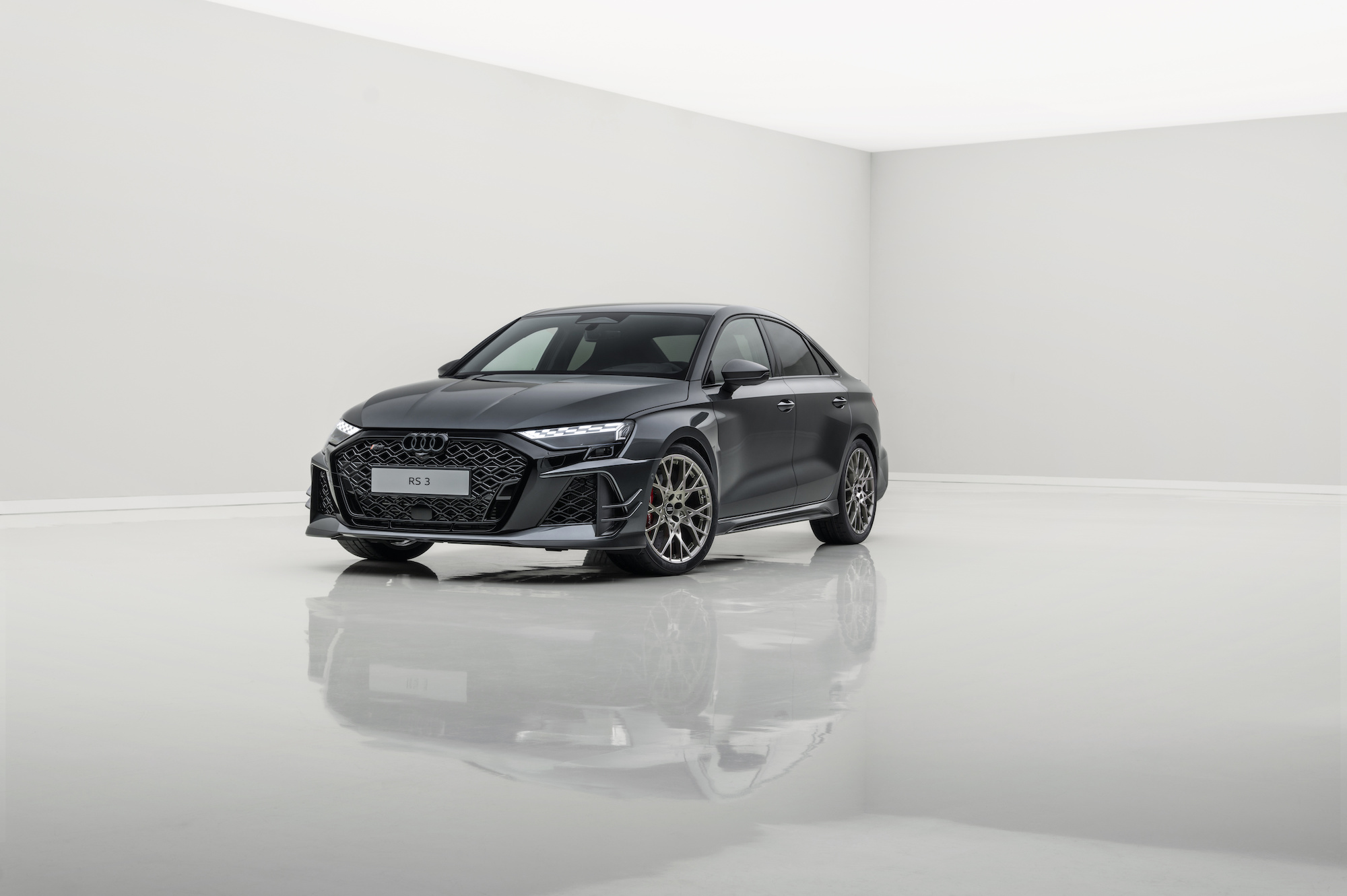 A sleek black Audi RS3 sports sedan showcased in a minimalist, bright studio setting with a reflective floor.