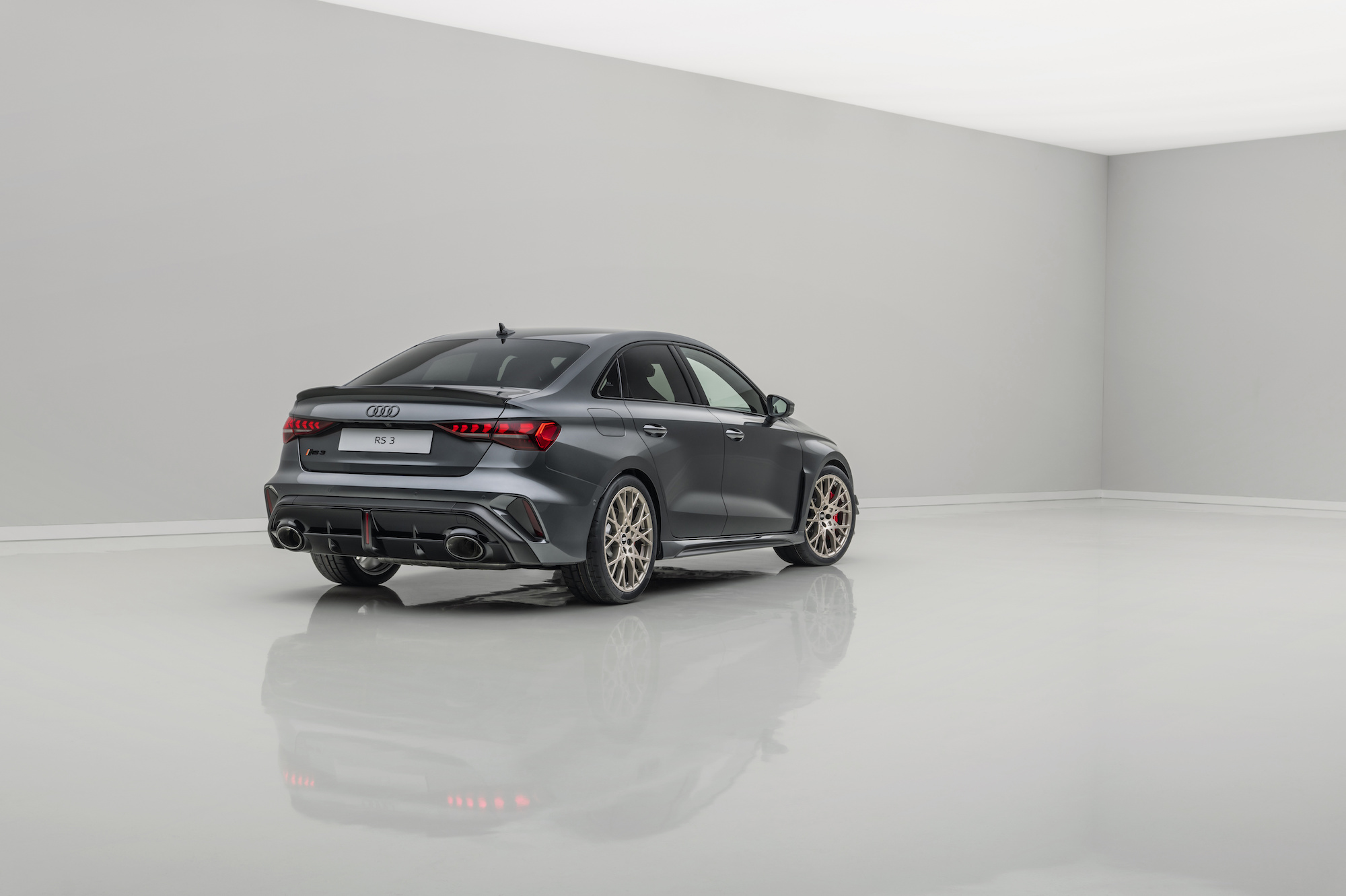 A sleek gray Audi RS 3 sports sedan, viewed from the rear, showcasing its aerodynamic design and distinctive taillights, against a minimalist white backdrop.