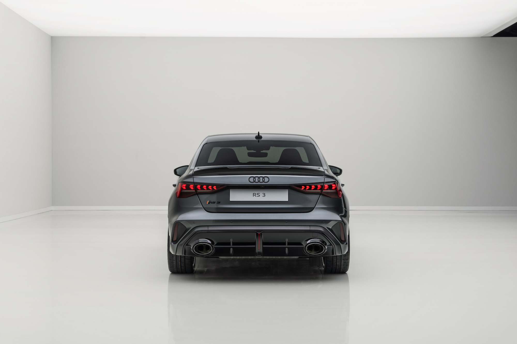 Rear view of a grey Audi RS 3 in a minimalistic display setting with a smooth white floor and wall.