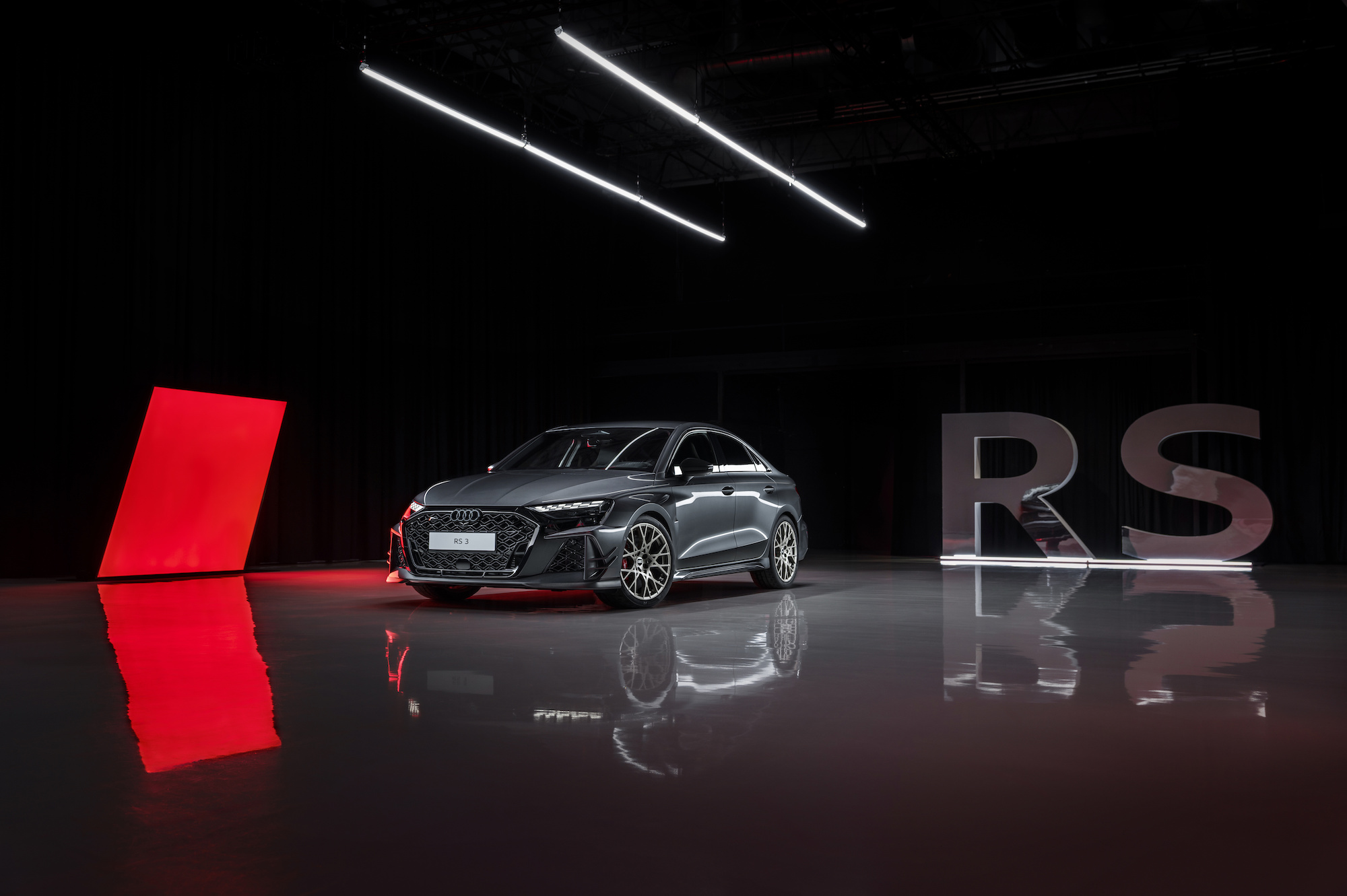 A sleek, gray Audi RS3 sports car displayed in a modern studio with dramatic lighting. A bright red geometric shape and translucent letters spelling 'RS' enhance the eye-catching presentation.