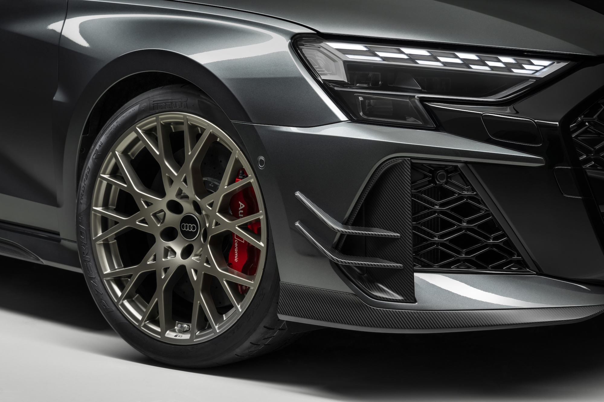 Close-up of a gray luxury car featuring a detailed alloy wheel with a unique spoke design and red brake calipers, showcasing the carbon fiber front splitter and aggressive front fascia.