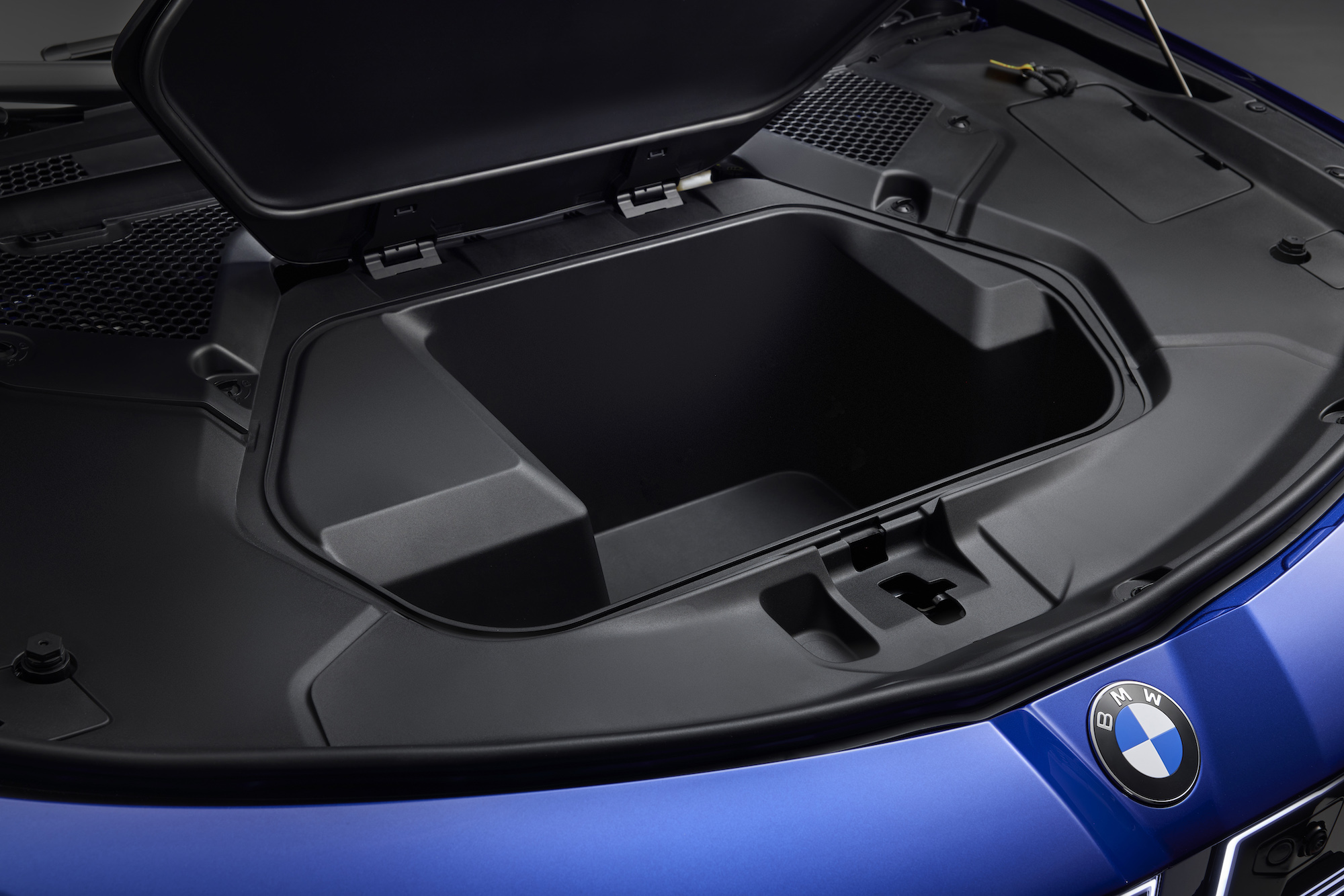 Close-up view of a car's engine compartment with the lid partially open, revealing a storage space and the BMW logo visible.