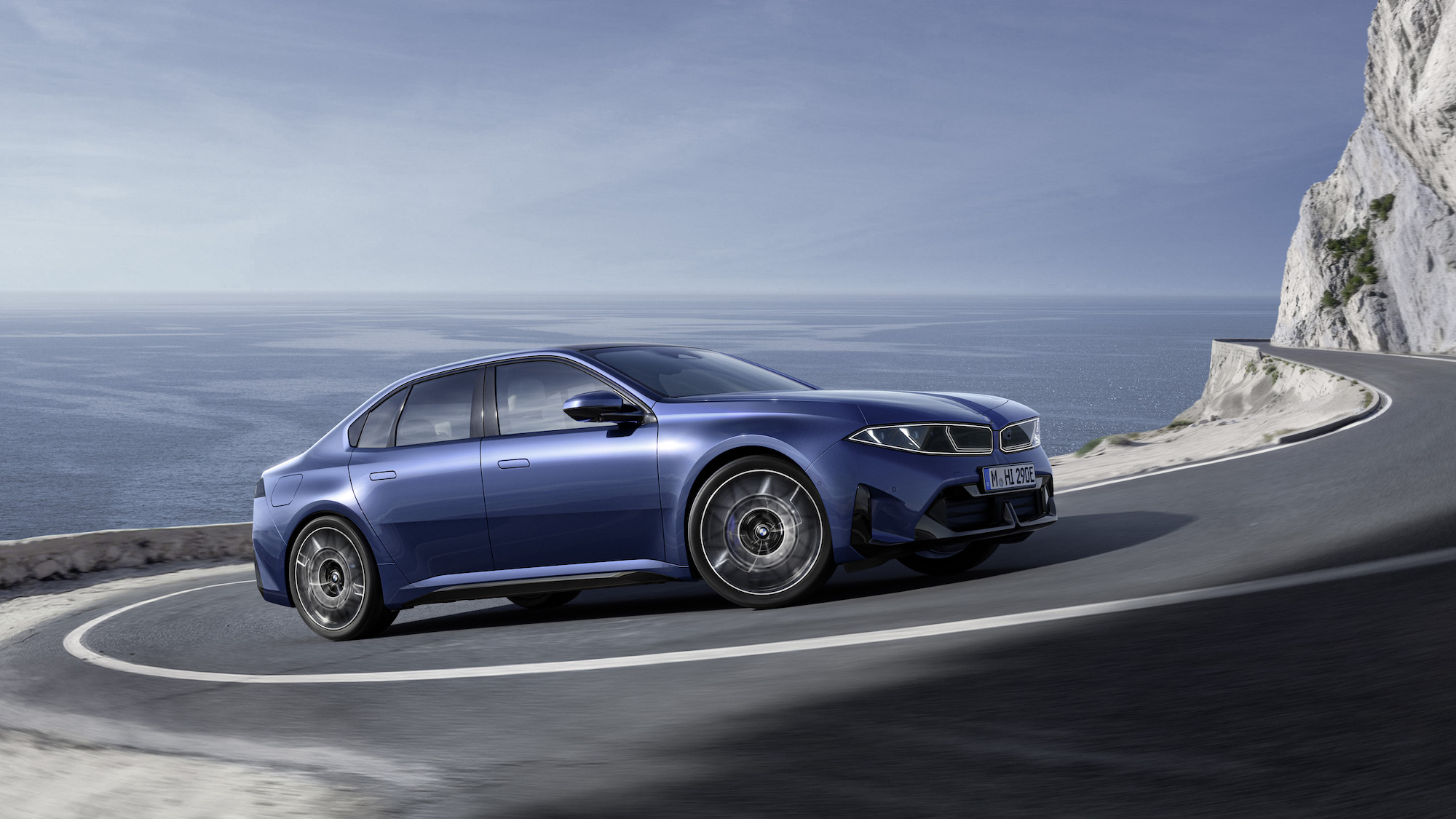 A blue sports sedan driving on a curved coastal road with a view of the ocean in the background.