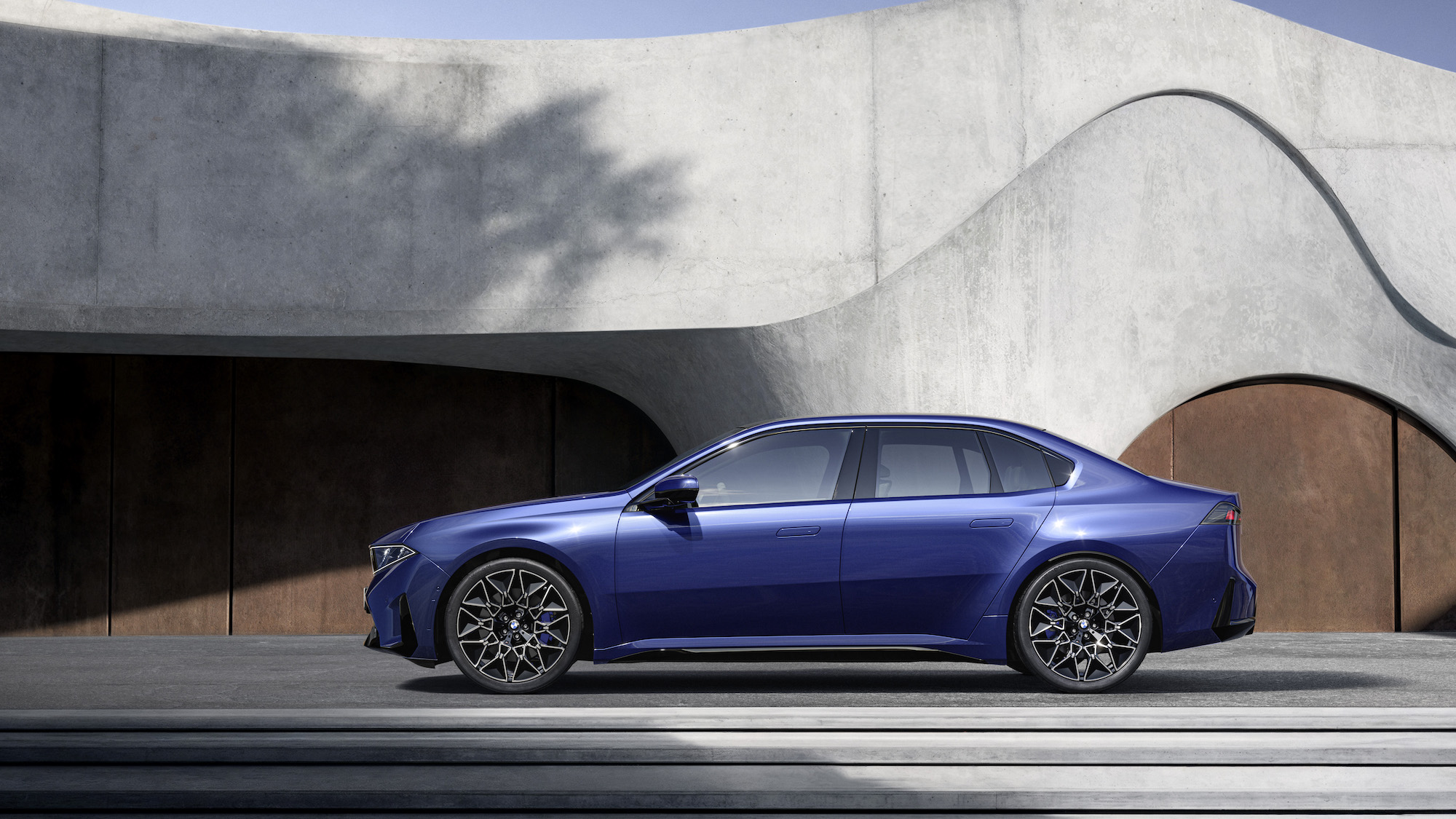 A blue luxury sedan parked in front of a modern architectural structure with curved concrete elements.
