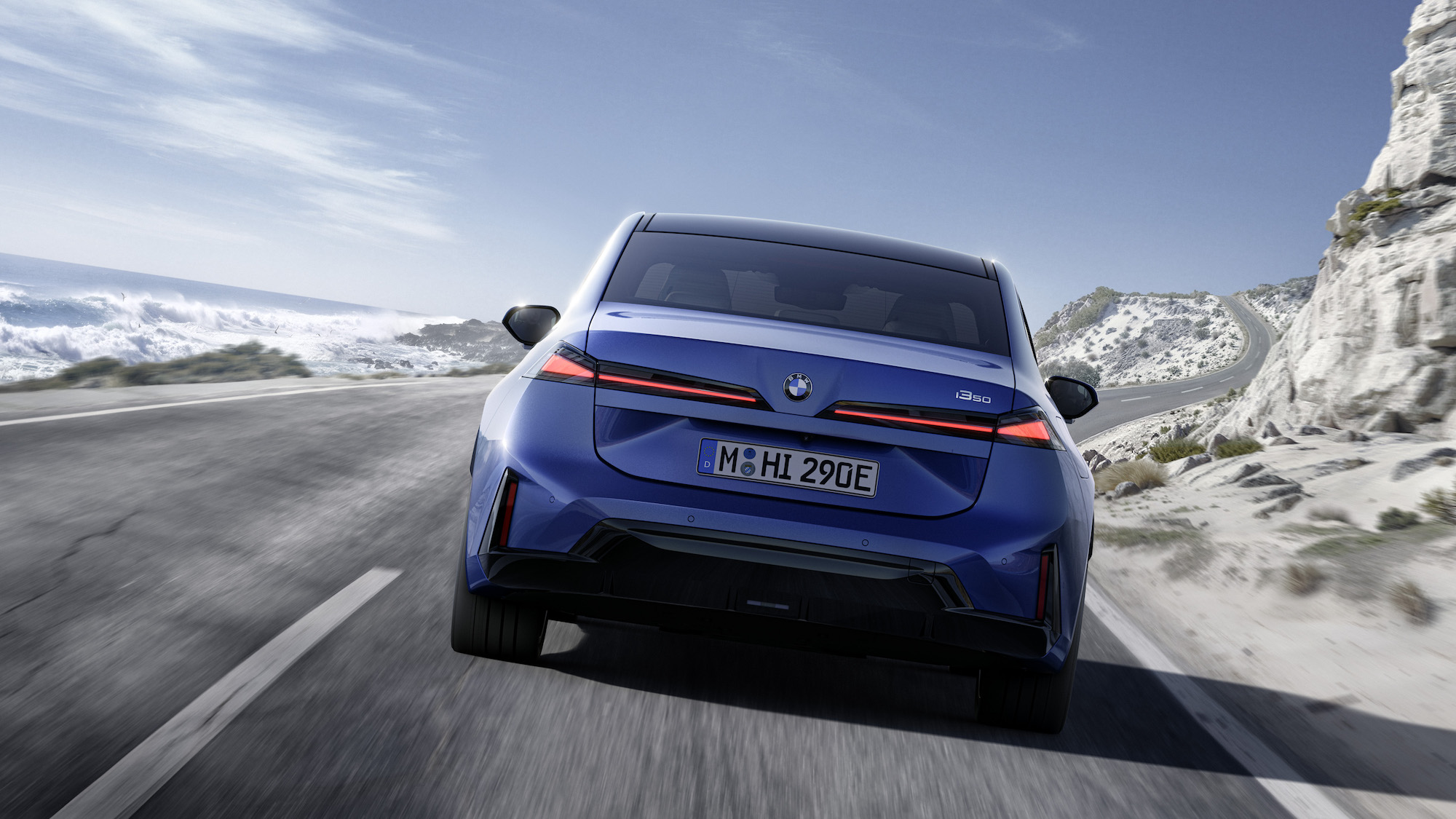 A blue BMW i3 30 driving along a coastal road with waves crashing in the background and a mountainous landscape on the side.