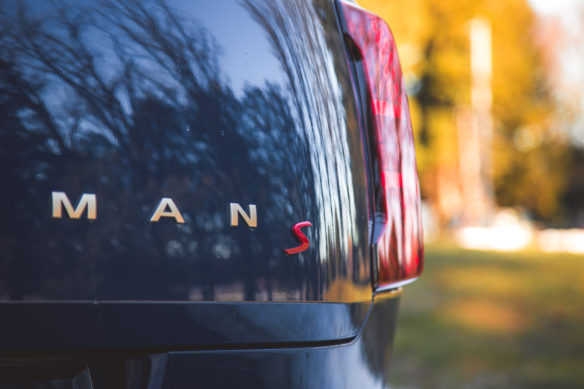 Close-up of the rear of a dark car displaying the logo letters 'MANS' and a red 'S', with a blurred background of trees and autumn colors.