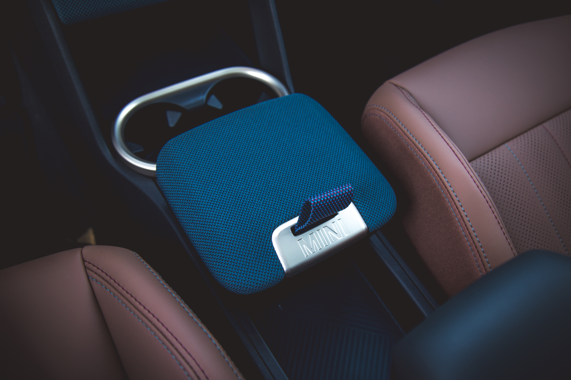 Close-up view of a blue textured armrest with a MINI logo, placed between two brown leather seats in a car interior.