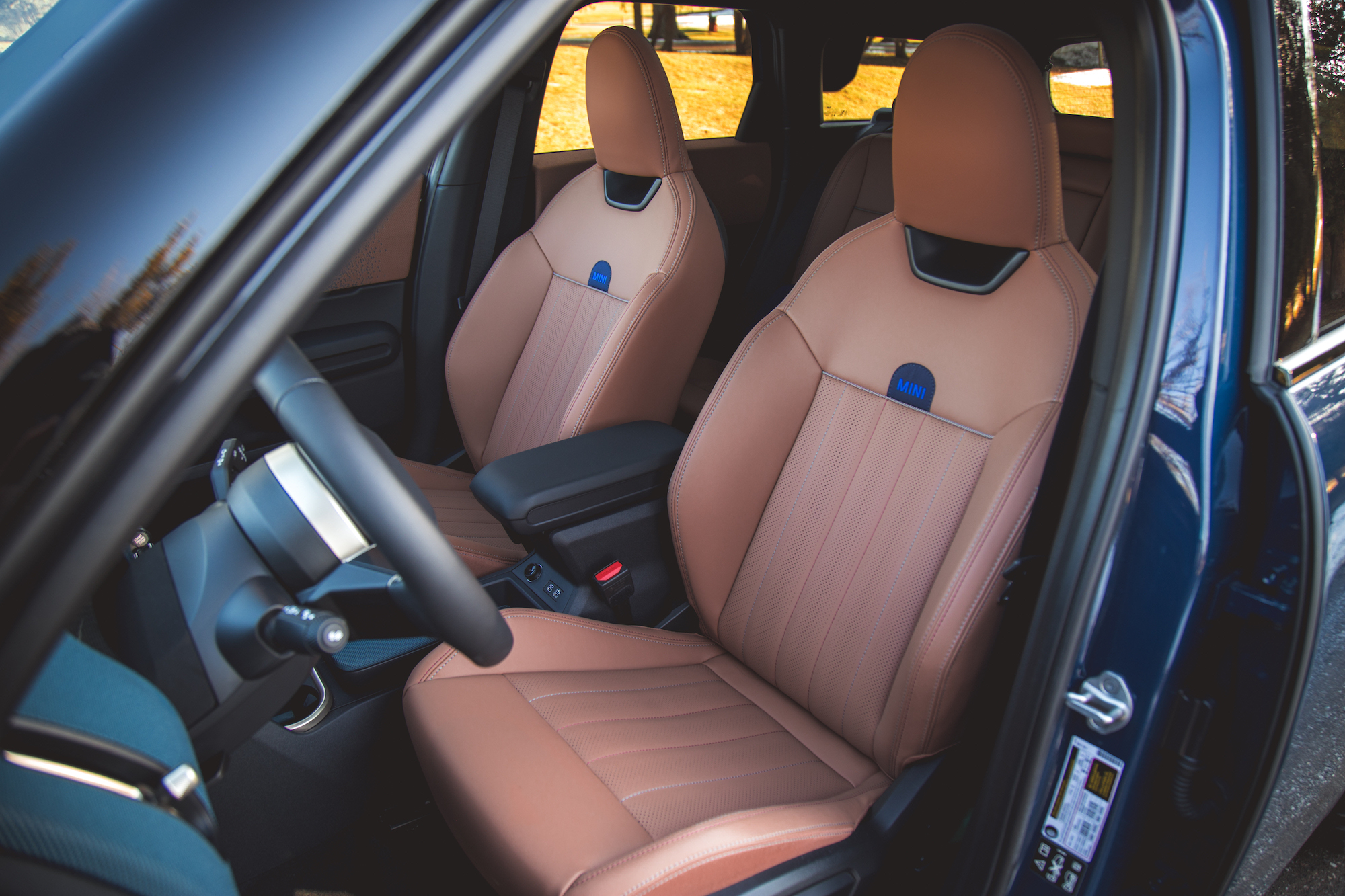 Interior view of a car featuring two tan leather seats with horizontal stitching and a central armrest, alongside a steering wheel.