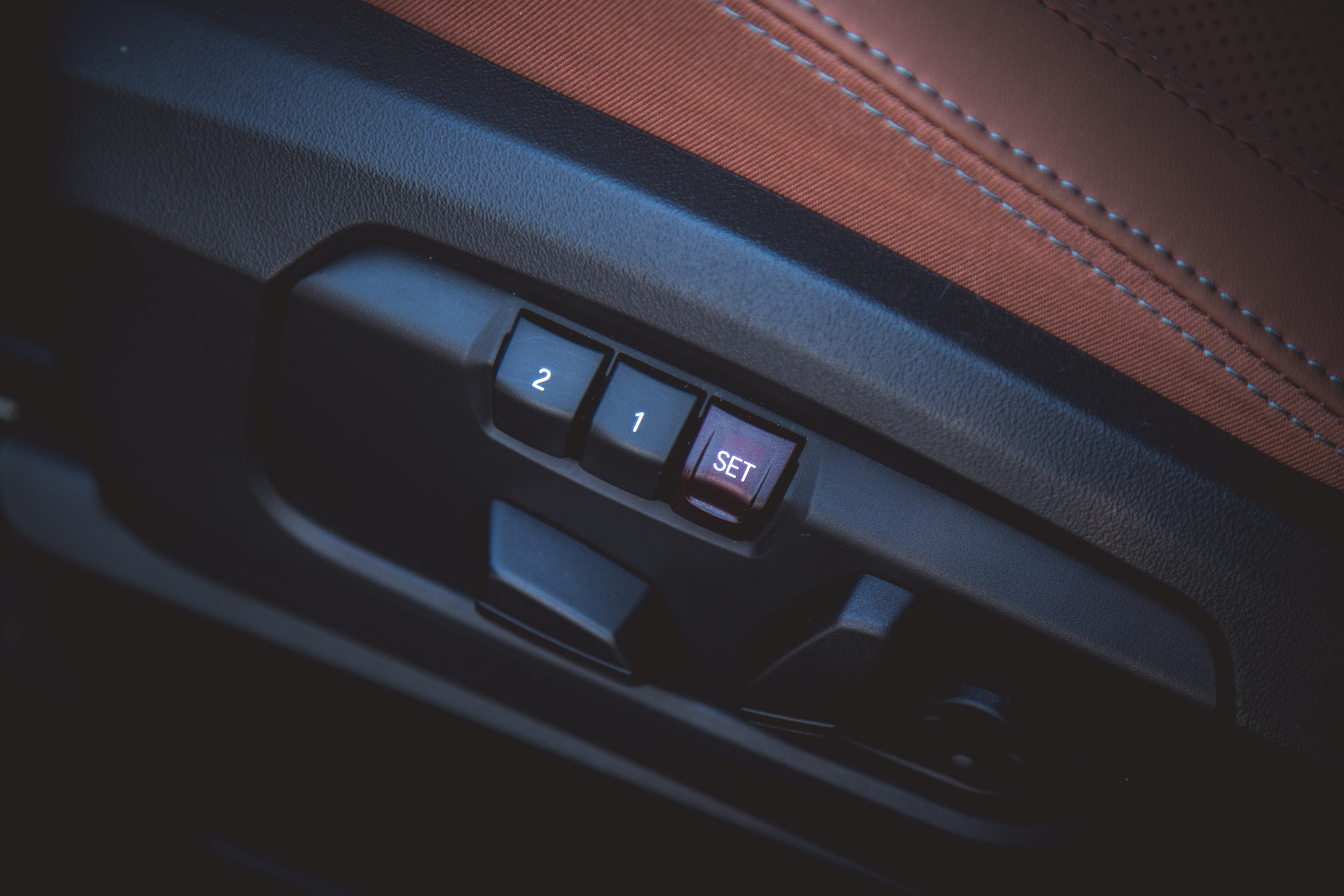 Close-up of a car seat adjustment control panel featuring buttons labeled '1', '2', and 'SET'. The panel is positioned against a brown leather seat cover.
