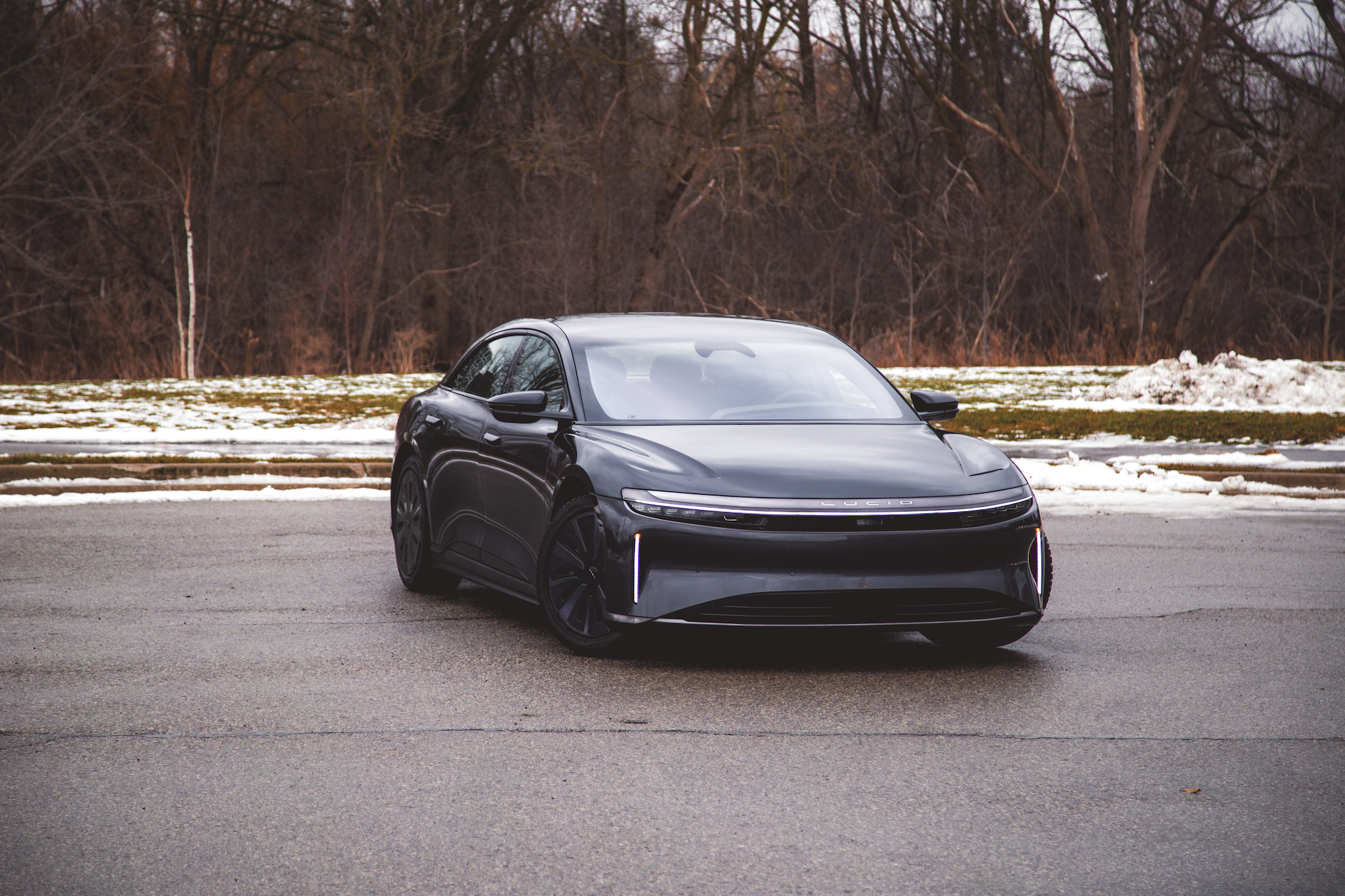 Un elegante coche eléctrico negro estacionado sobre una superficie de asfalto gris con un paisaje nevado y árboles desnudos al fondo.
