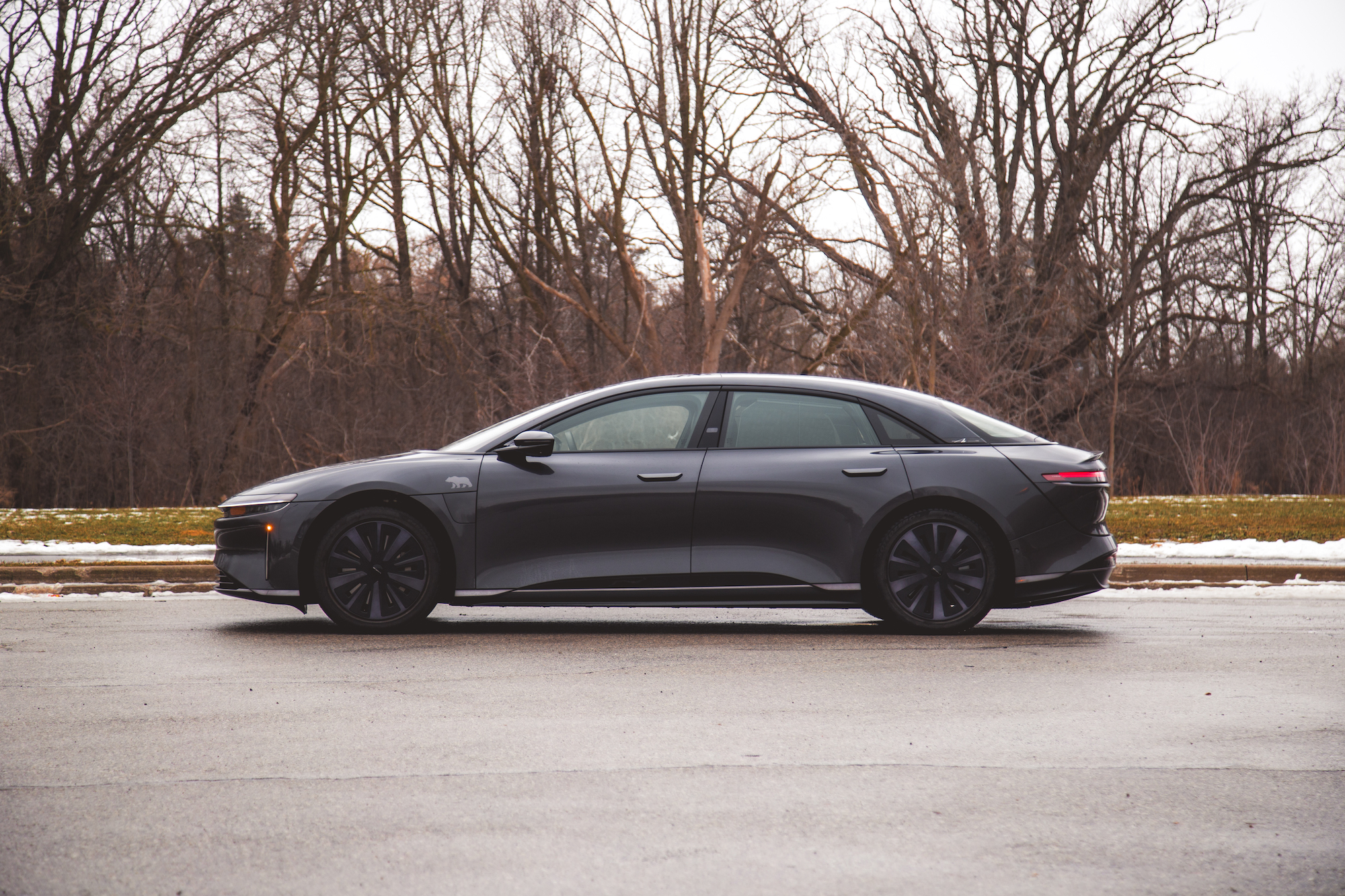 Vista lateral de un moderno sedán eléctrico gris estacionado en una carretera con árboles desnudos al fondo.