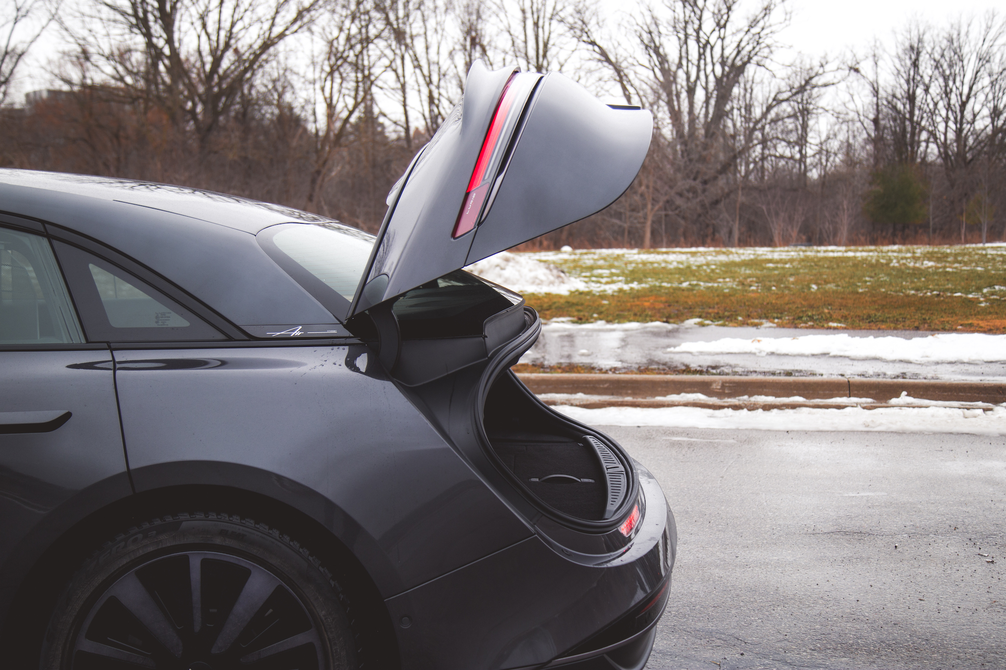Un coche negro con el maletero abierto, frente a un paisaje invernal con árboles desnudos y manchas de nieve.