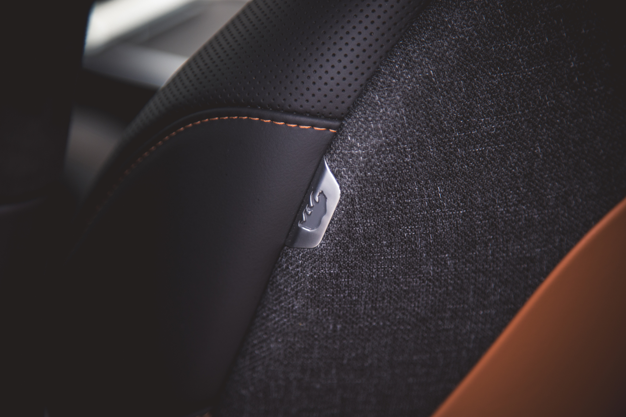Primer plano de un asiento para automóvil con una mezcla de cuero negro y tela gris, con costuras naranjas y un detalle de logotipo metálico.