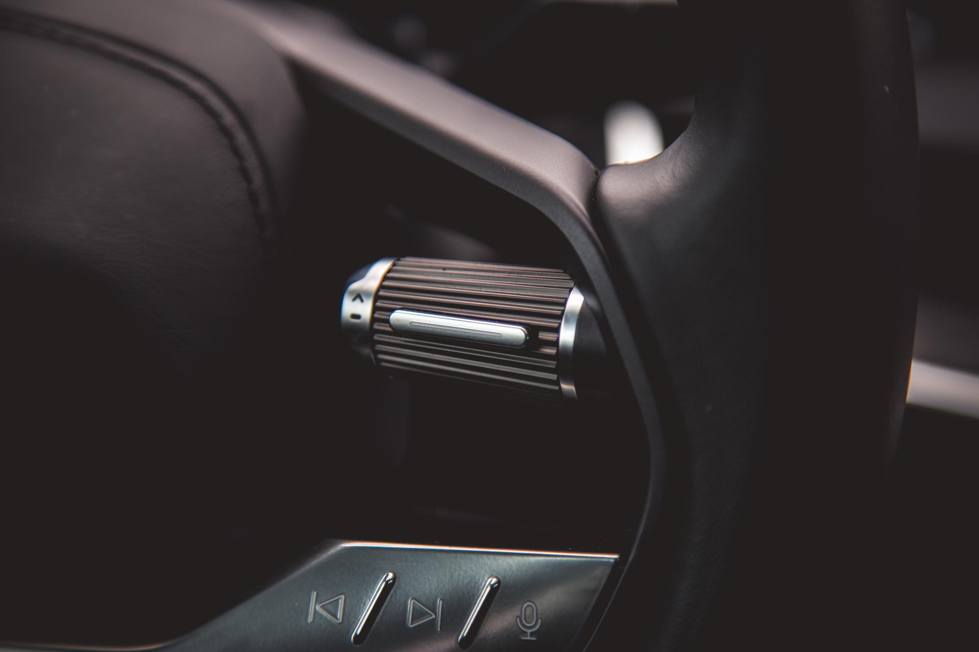 Primer plano de la perilla de control de un vehículo con agarre texturizado y detalles plateados, con botones de navegación y control por voz cercanos.