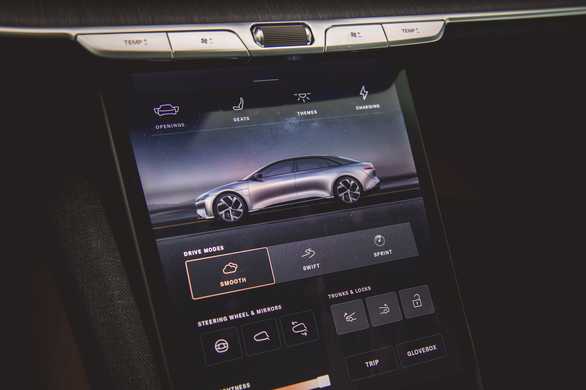 Primer plano de la interfaz de pantalla táctil de un automóvil moderno que muestra configuraciones para modos de manejo, asientos y opciones de carga, con un elegante gráfico plateado del automóvil.